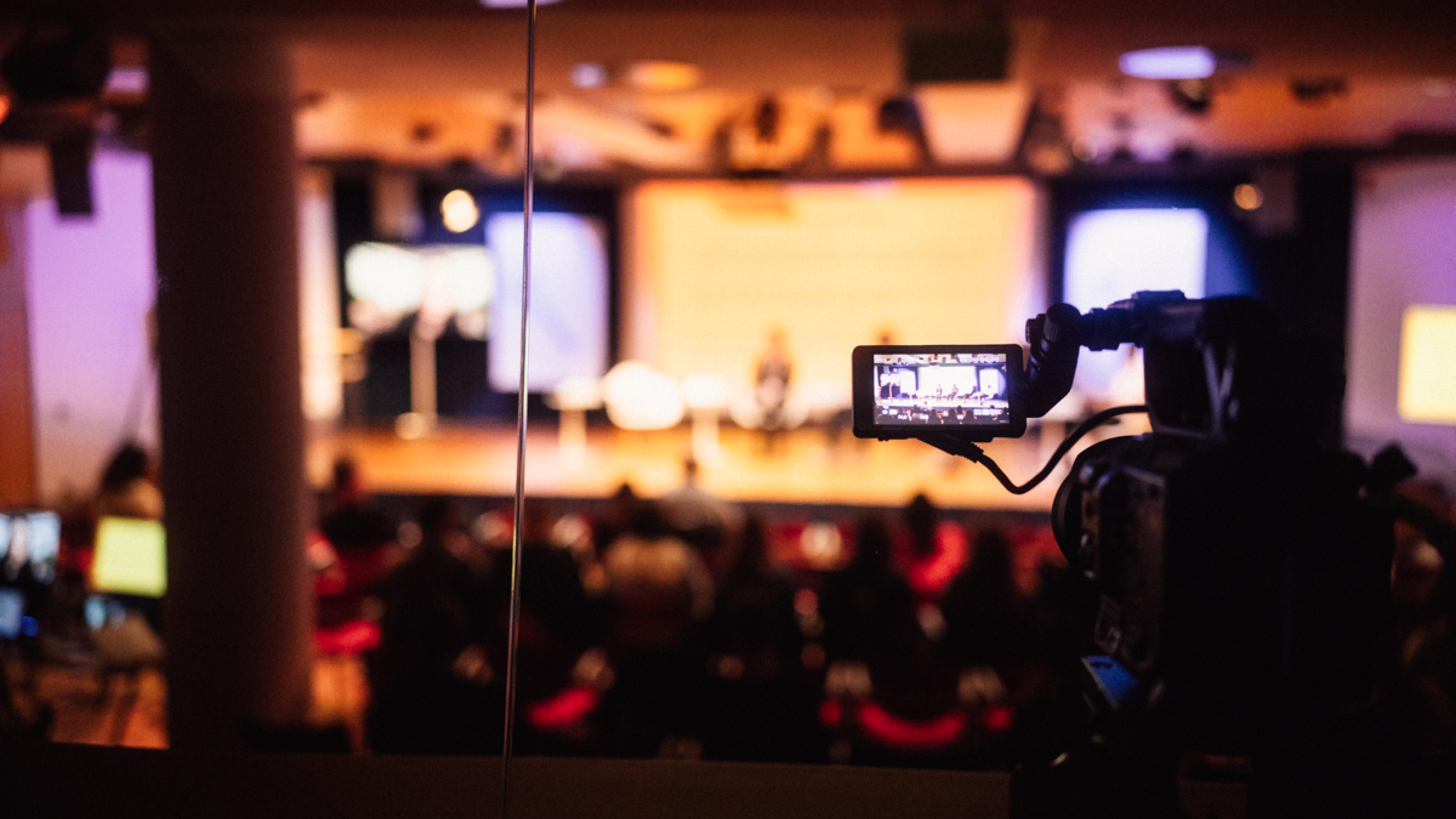 A camera monitor shows a panel of four persons on the podium.