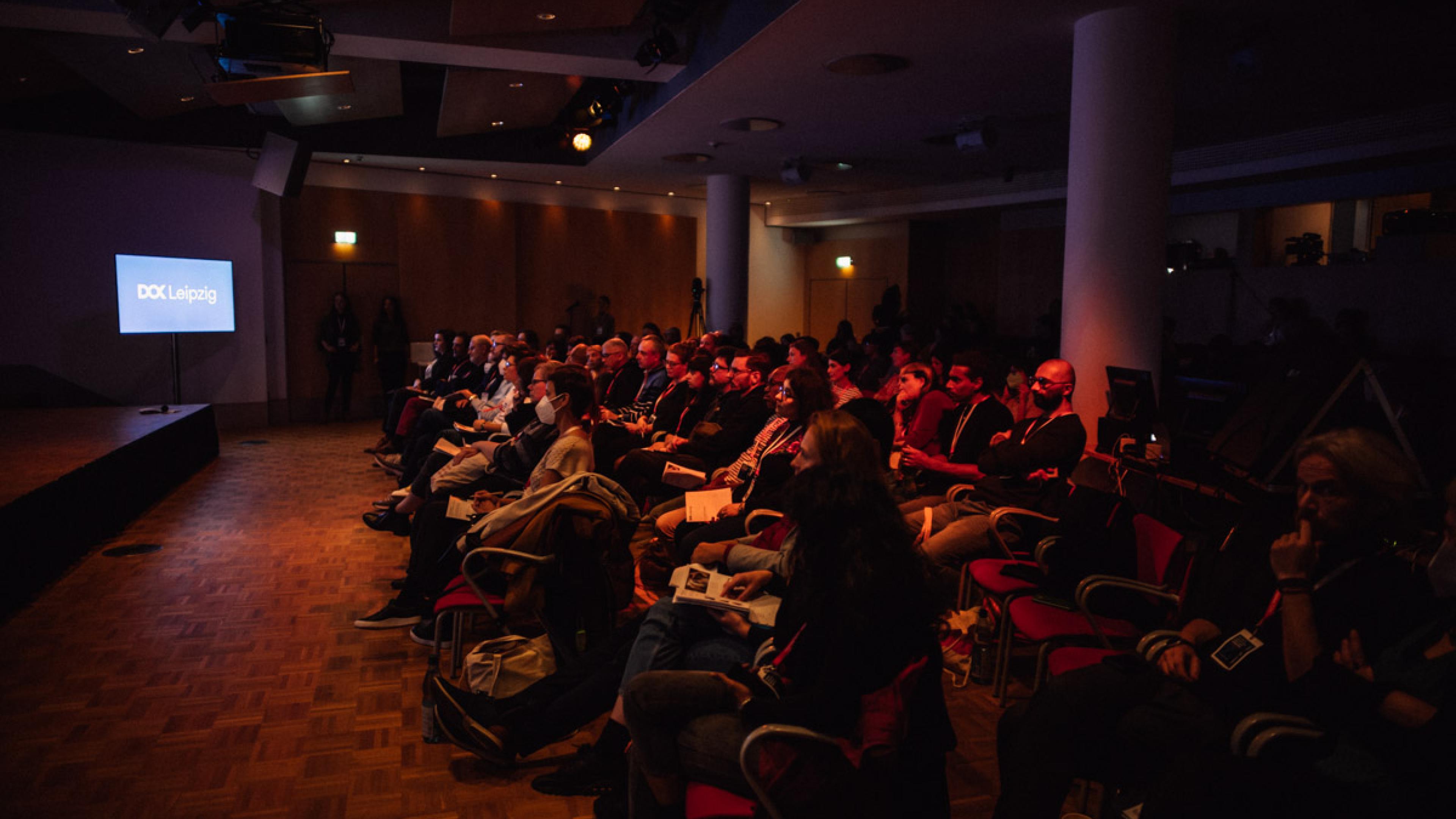 Audience in a conference room. A screen at the side shows the DOK Leipzig logo.