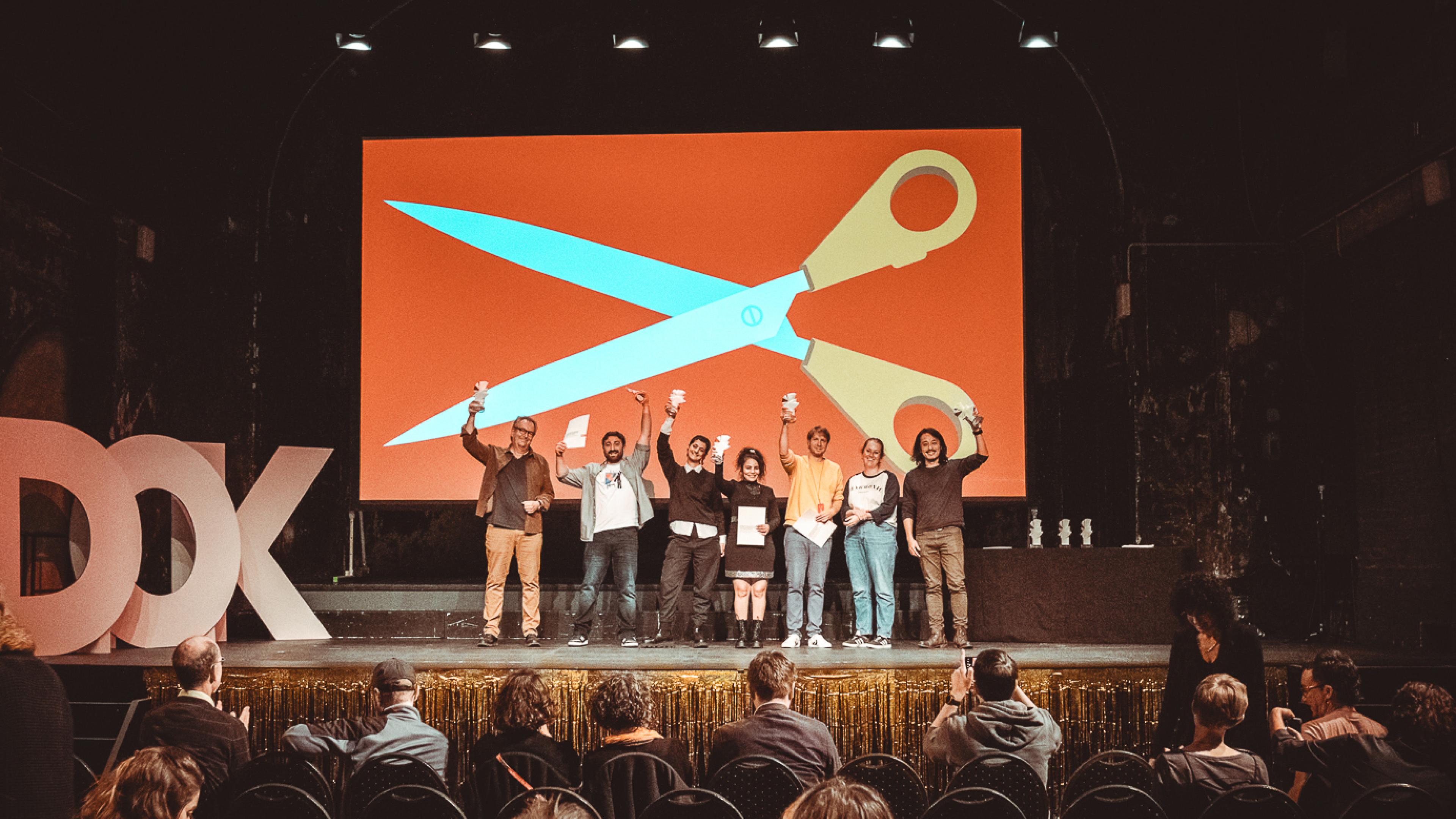 Sieben Personen stehen auf einer Bühne und halten Preisstatuetten in die Höhe. Im Hintergrund das Festivalmotiv von DOK Leipzig 2023: eine riesige Schere.