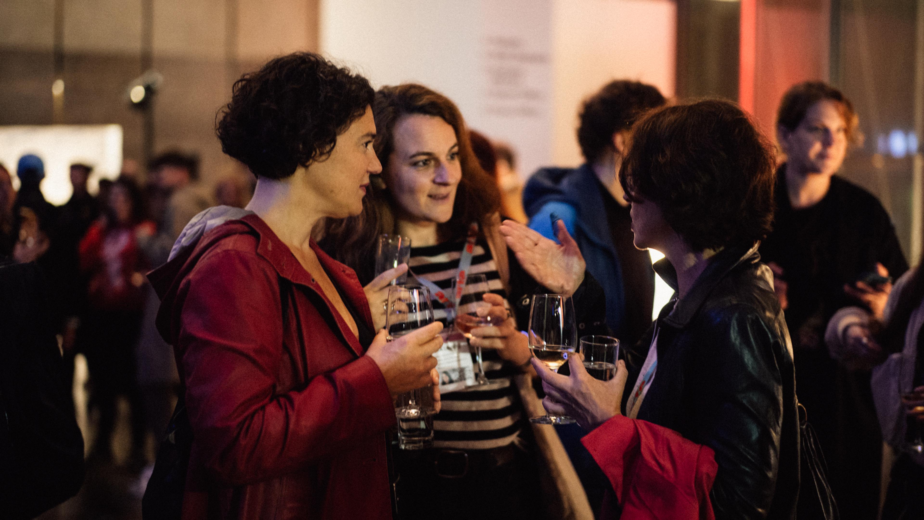 Three women stand together, holding glasses in their hands, and chat. Around them a lot of other persons.