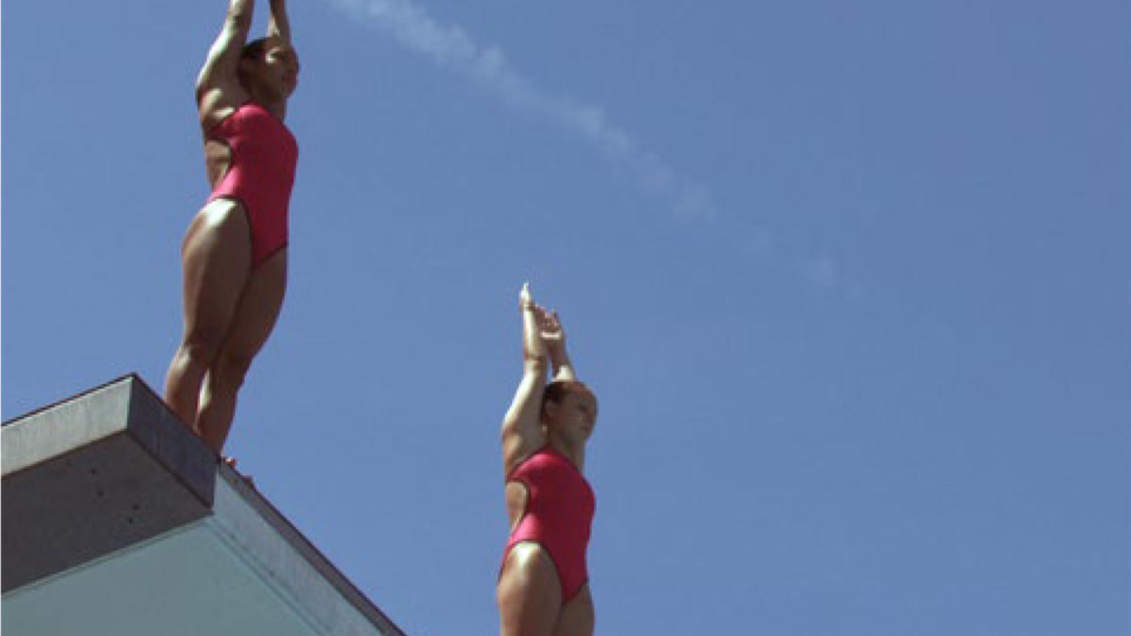 Zwei Frauen in roten Badeanzügen stehen bereit zum Absprung auf einem Sprungturm.