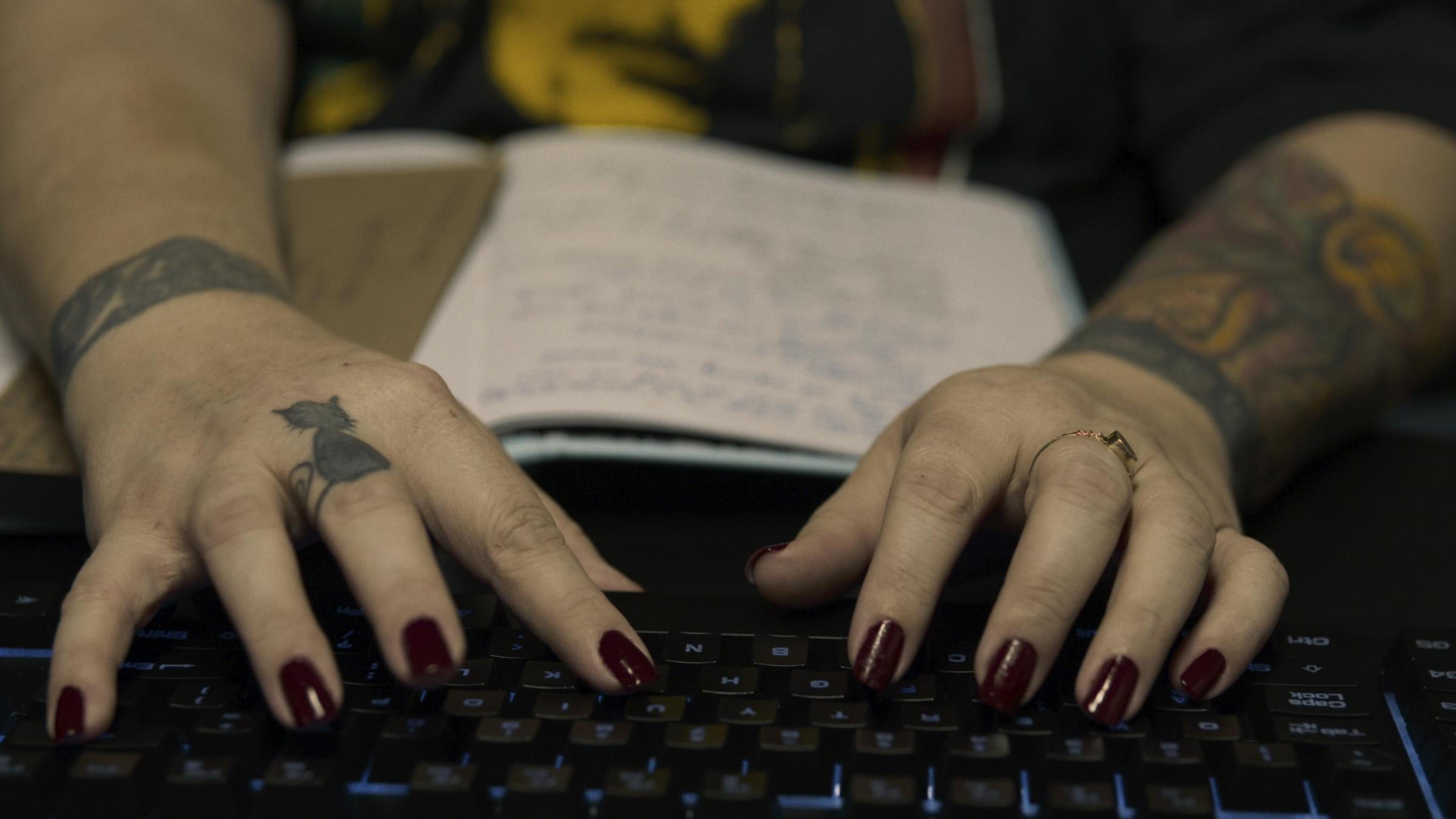 Zwei tätowierte Hände mit dunkel lackierten Fingernägeln tippen auf einer Computertastatur.