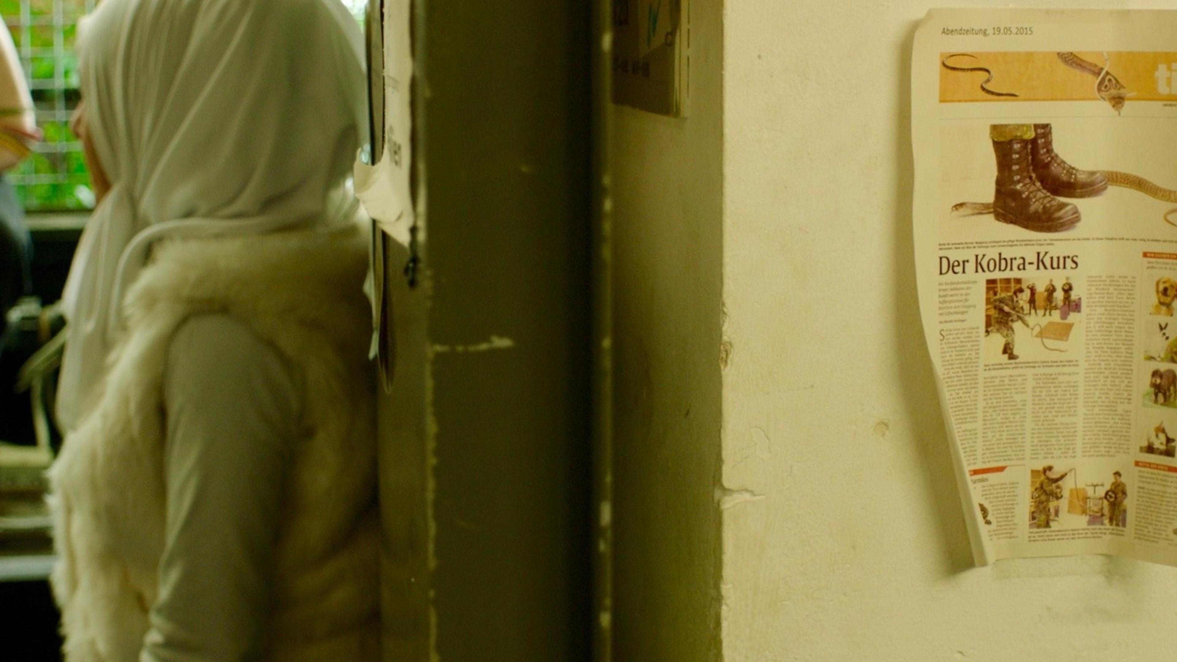 Eine Frau mit Kopftuch lehnt an einer Wand in einem Flur. Neben ihr hängt ein Zeitungsausschnitt über einen Kobra-Kurs.