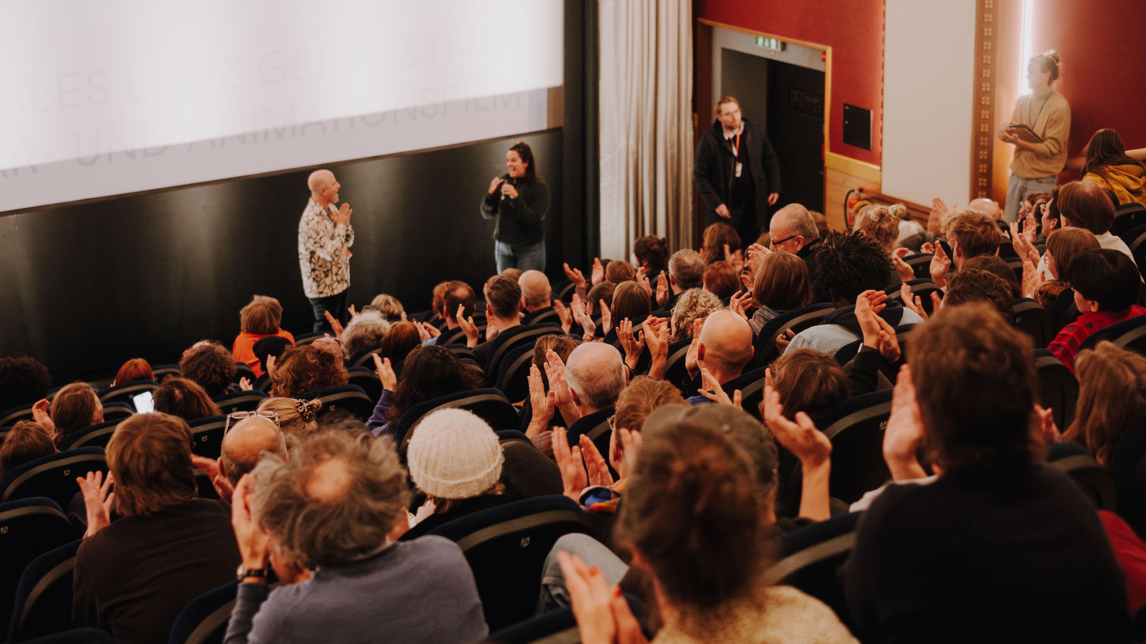 Ein voller Kinosaal, zwei Menschen stehen vor der Leinwand und sprechen miteinander, die Menge applaudiert