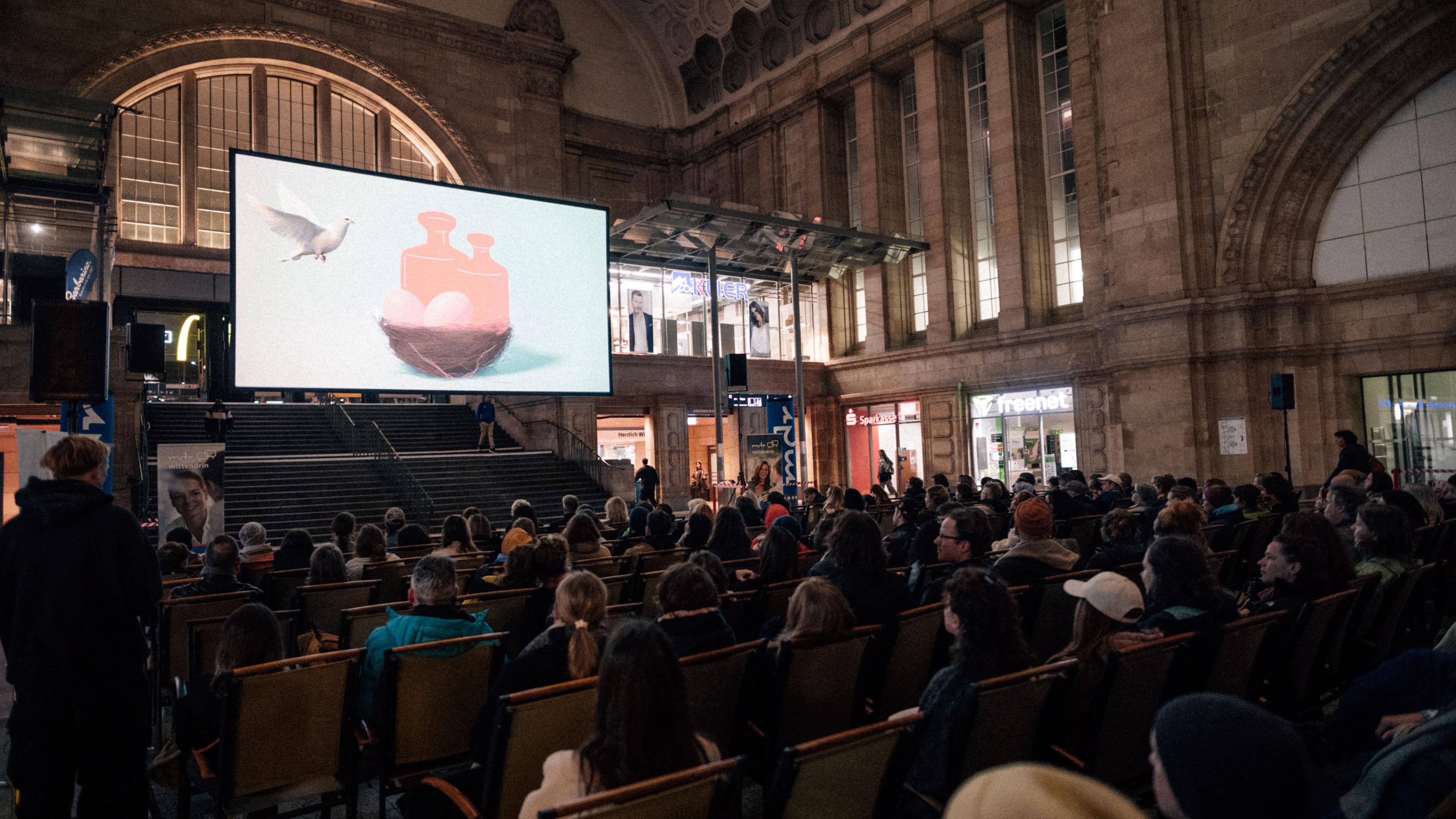 In der Osthalle vom Hauptbahnhof ist eine Kinoleinwand aufgebaut, viele Menschen sitzen auf Stühlen und schauen einen Trailer, in dem eine animiert Taube zu ihrem Nest voller Eier und Gewichten fliegt.