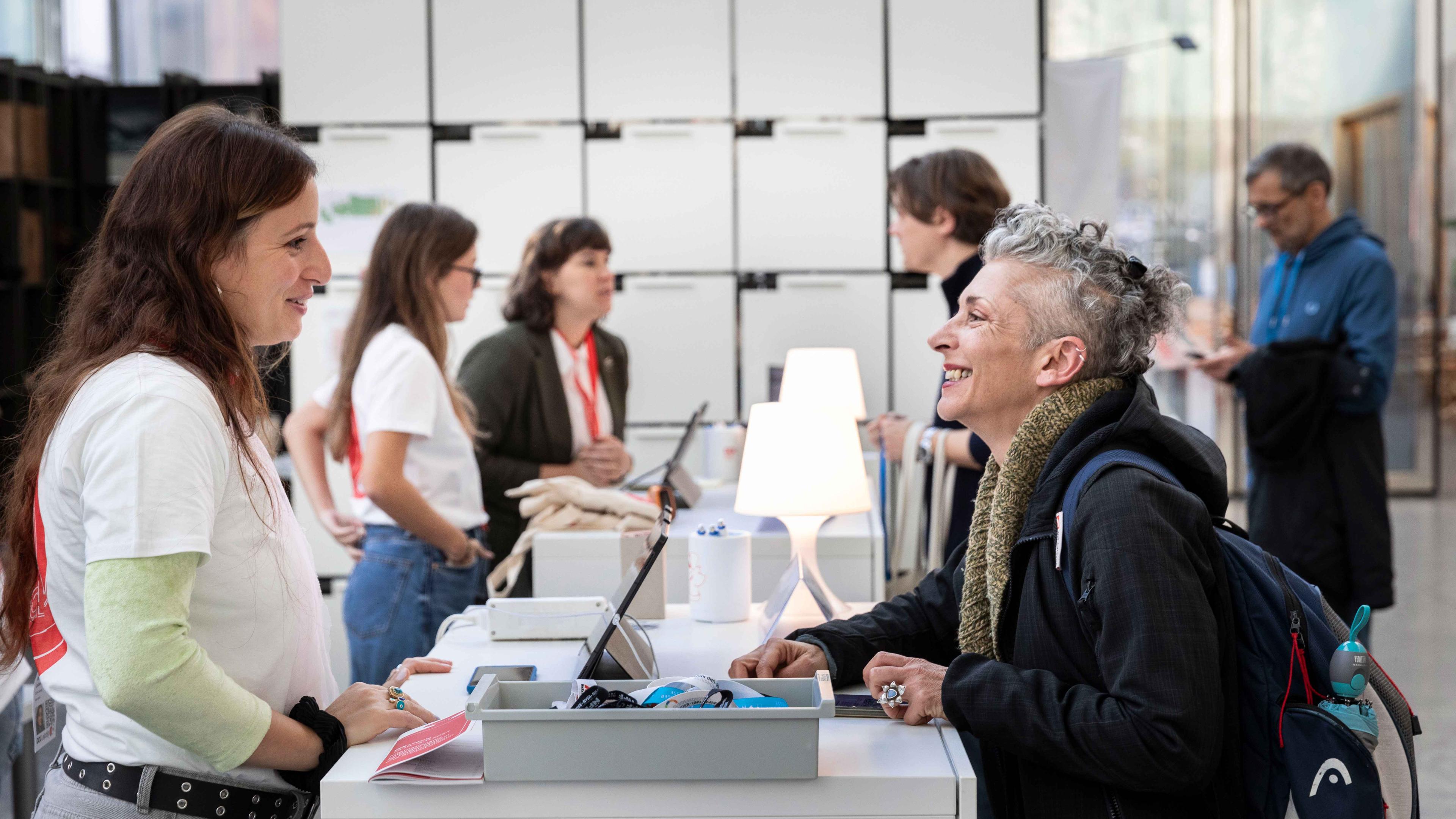 Am Gästecounter lachen sich eine Festivalgästin und eine Volunteerperson bei der Abholung der Akkreditierung fröhlich an