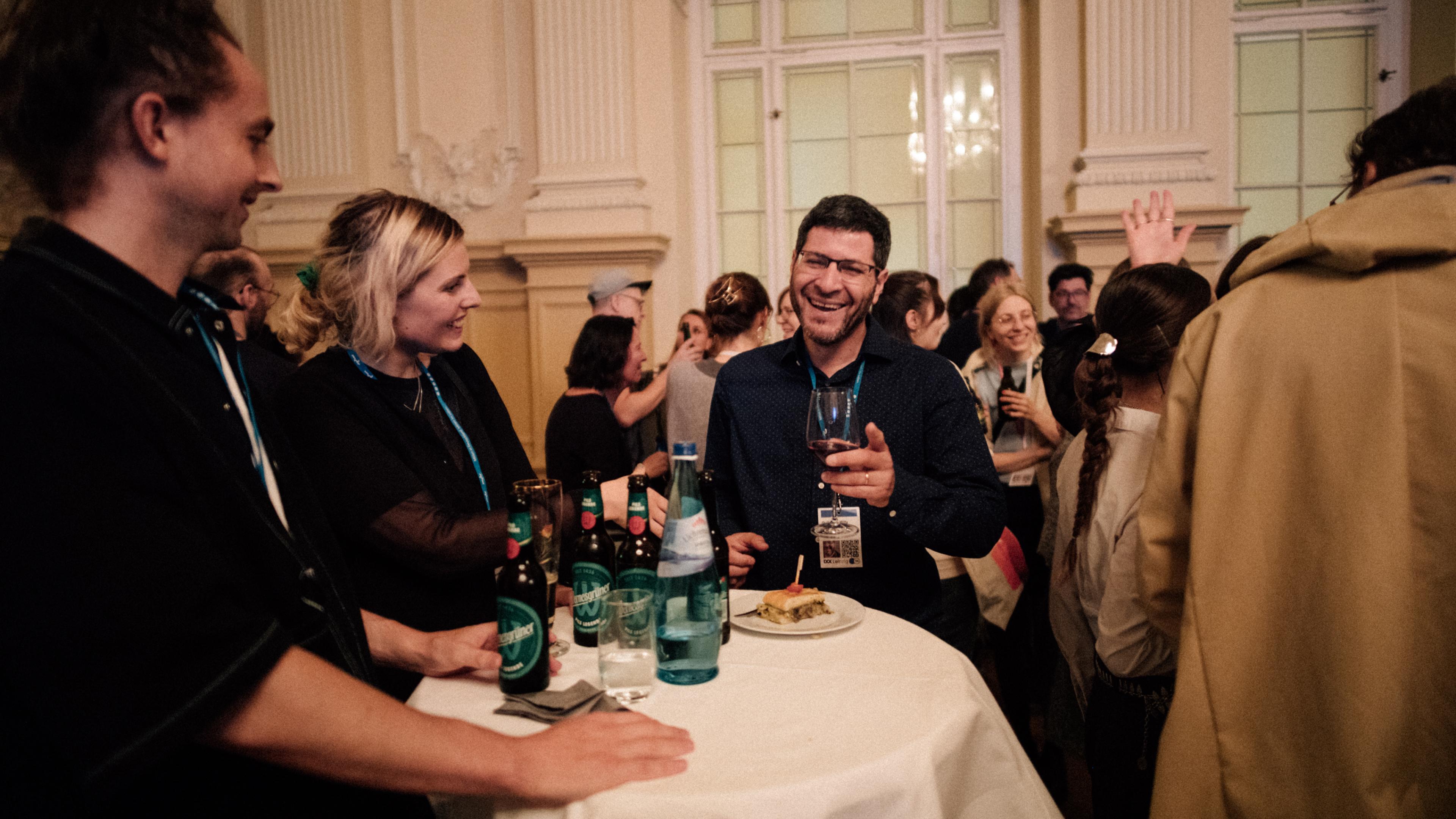 Eine kleine Gruppe Menschen stehen um einen Tisch herum, trinken aus Flaschen und Weingläsern und sehen gut gelaunt aus