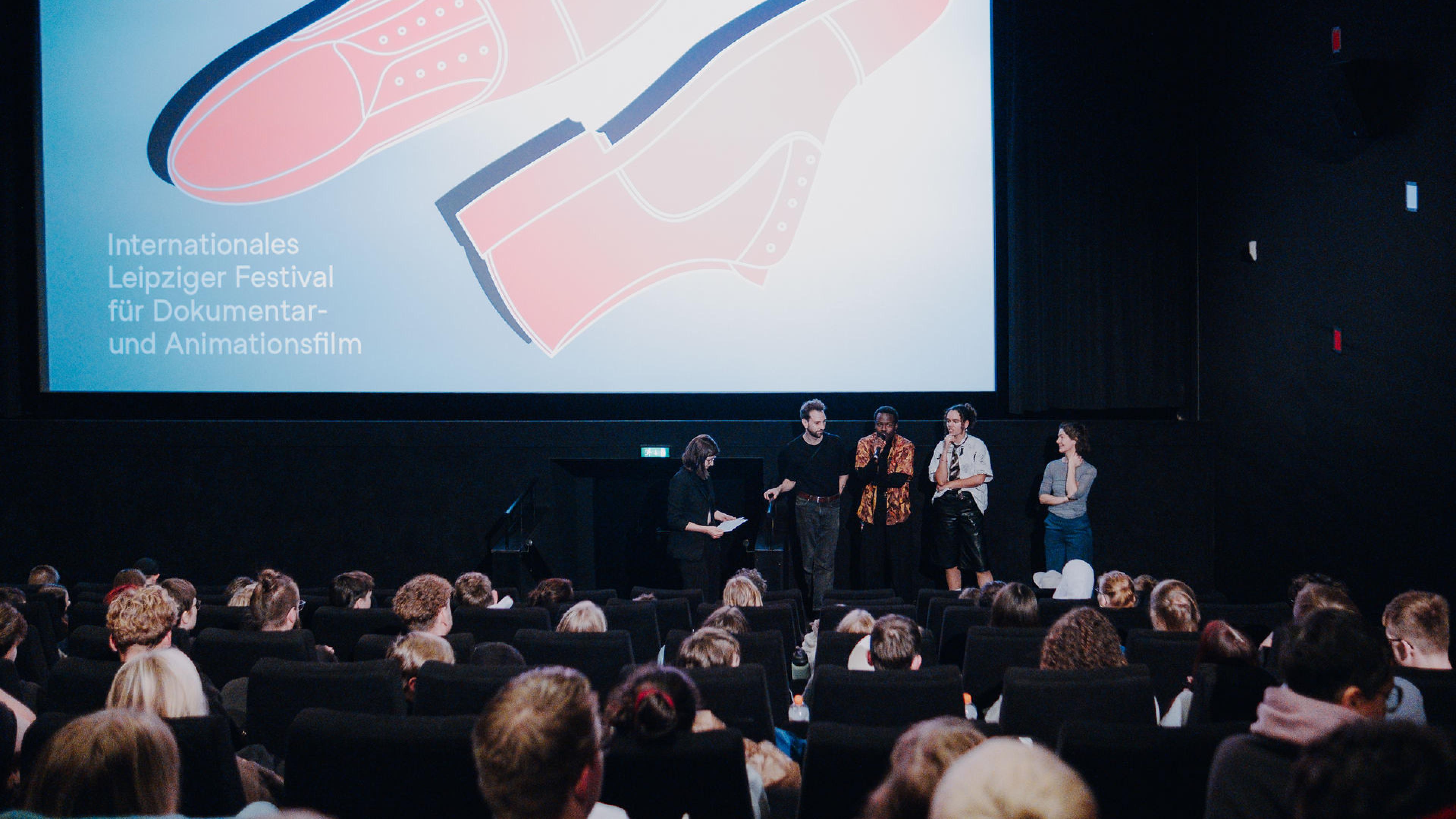 Ein voller Kinosaal mit dem 2025 DOK Leipzig Motiv auf der Leinwand. Davor steht ein Filmteam im Gespräch