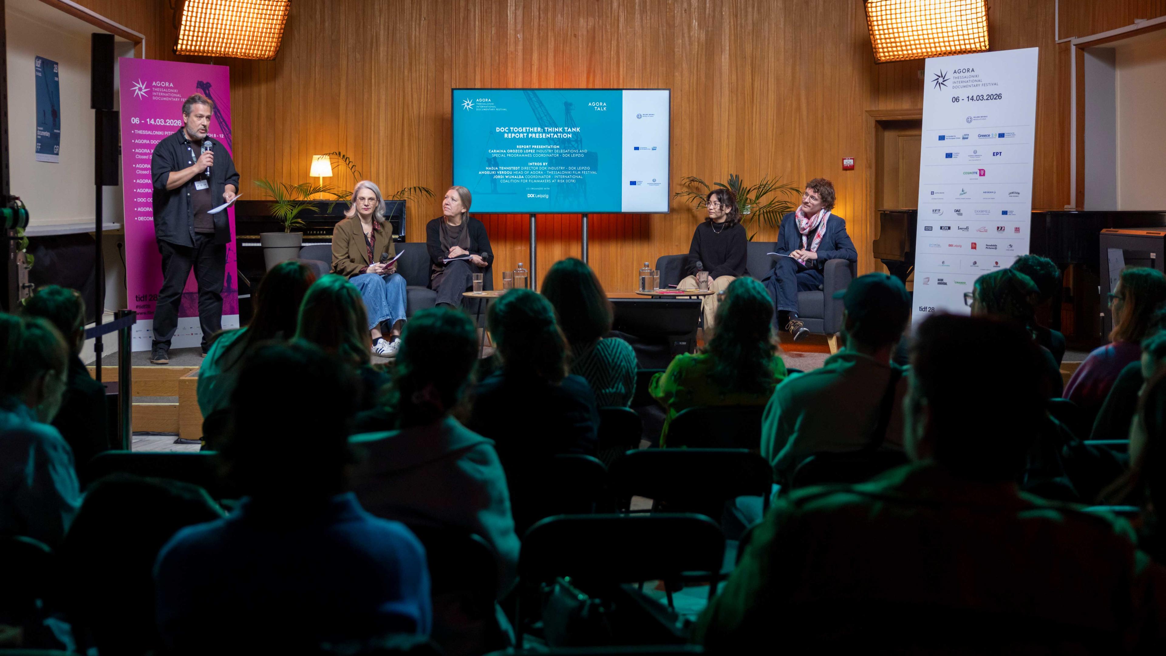 In einem überfüllten Raum sitzen fünf Personen auf dem Podium. Hinter ihnen befindet sich eine Leinwand mit der englischen Aufschrift: Doc Together Think Tank Report Presentation