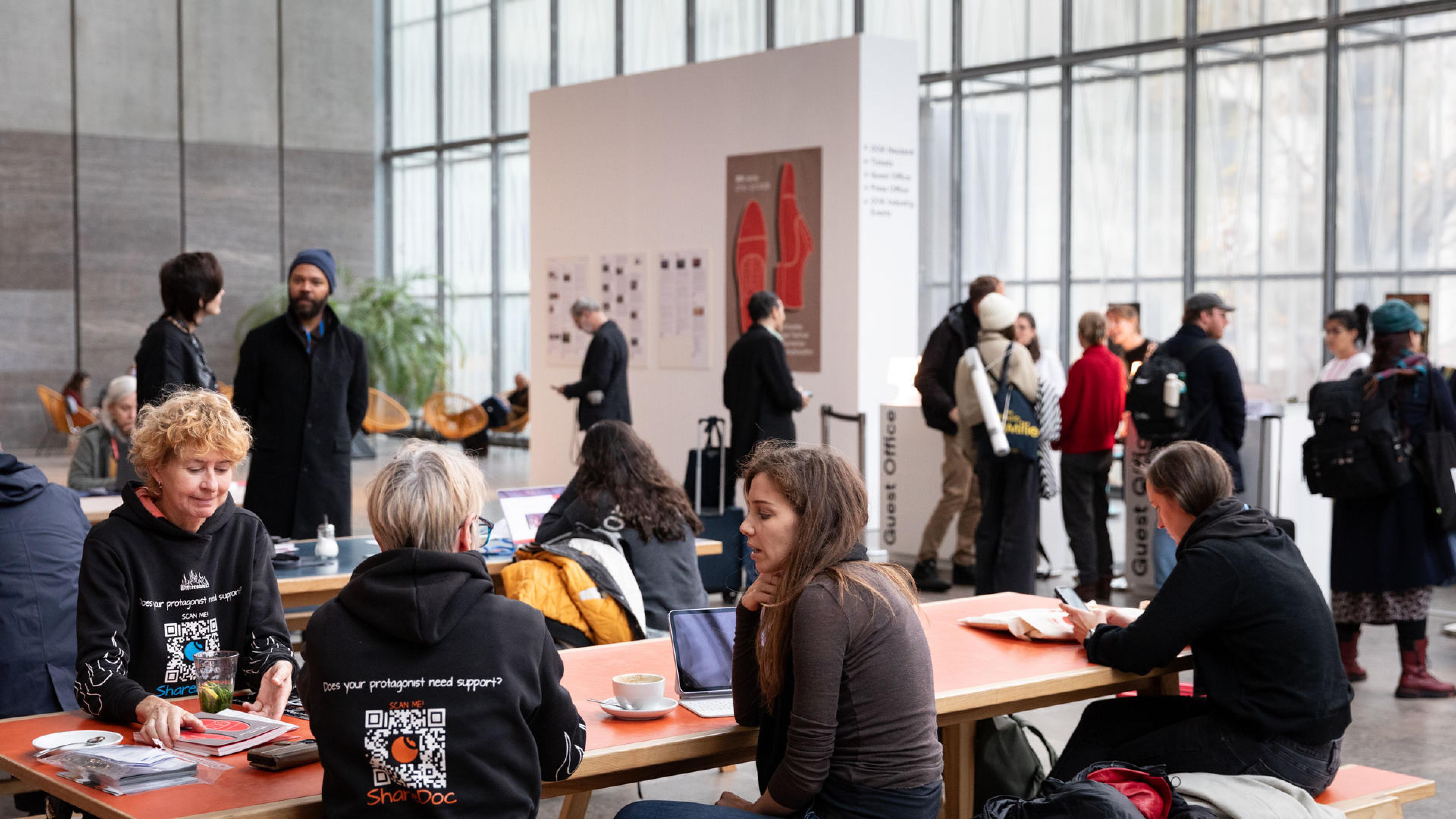 Große, helle Halle mit langen Tischen, an denen viele Personen sitzen, Kaffee trinken und sich unterhalten. Im Hintergrund ist Gästecounter zu sehen und daneben eine Wand mit Festivalplakat und ausgedruckten Presseberichten.