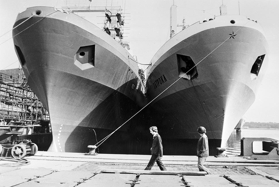 Zwei Personen gehen an der Kaimauer einer Werft entlang. Dort liegen zwei große Schiffe direkt nebeneinander auf einem Trockendeck. Sie erheben sich wie Riesen aus dem Wasser.