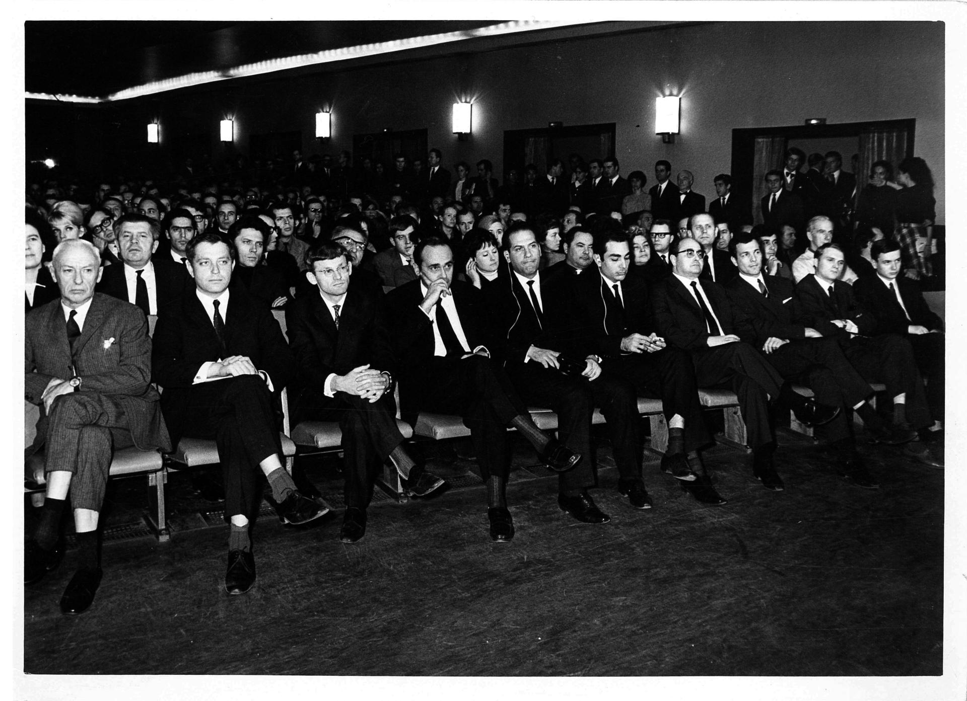 Schwarz-Weiß-Foto auf die ersten Reihen in einem voll besetzten Kinosaal. In der ersten Reihe sitzen ausschließlich Männer in dunklen Anzügen und weißen Hemden mit Krawatte.