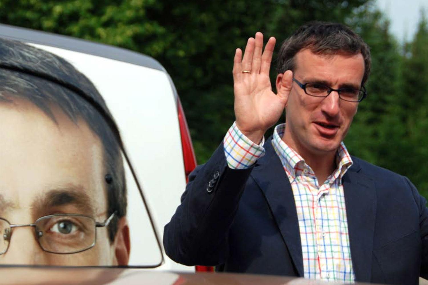 Ein Mann steht vor einem Wahlplakat mit einem Bild von sich selbst und winkt.