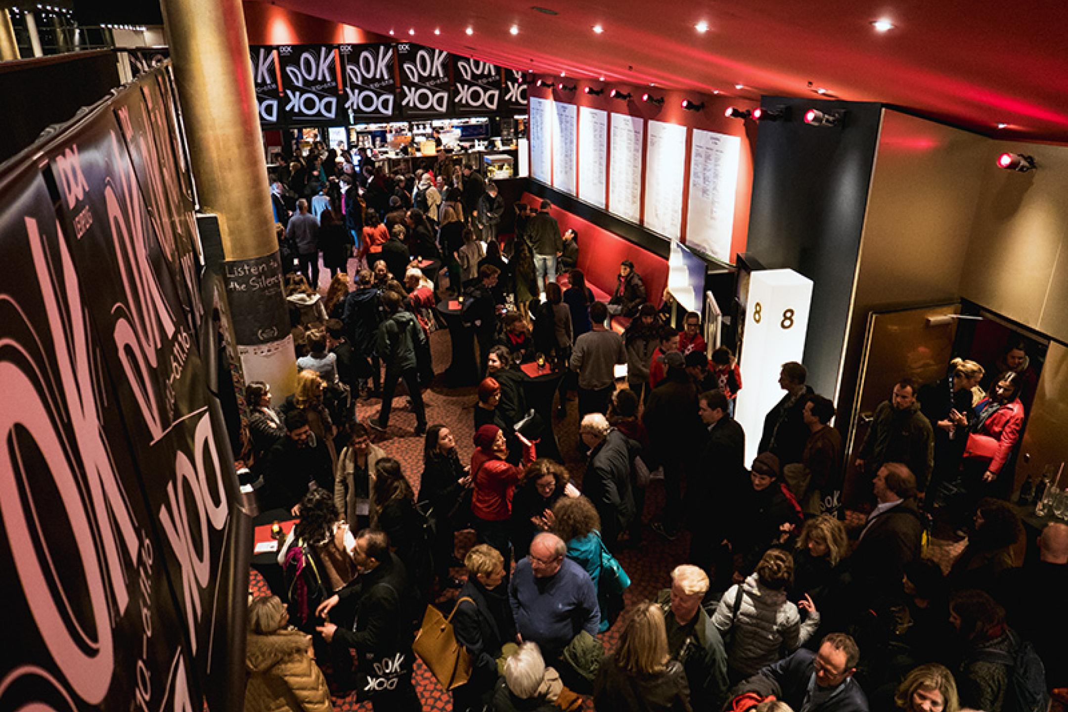 Foyer des Cinestar Leipzig mit vielen Besucherinnen und Besuchern, die ins Kino strömen, viele DOK Leipzig Plakate im Hintergrund.