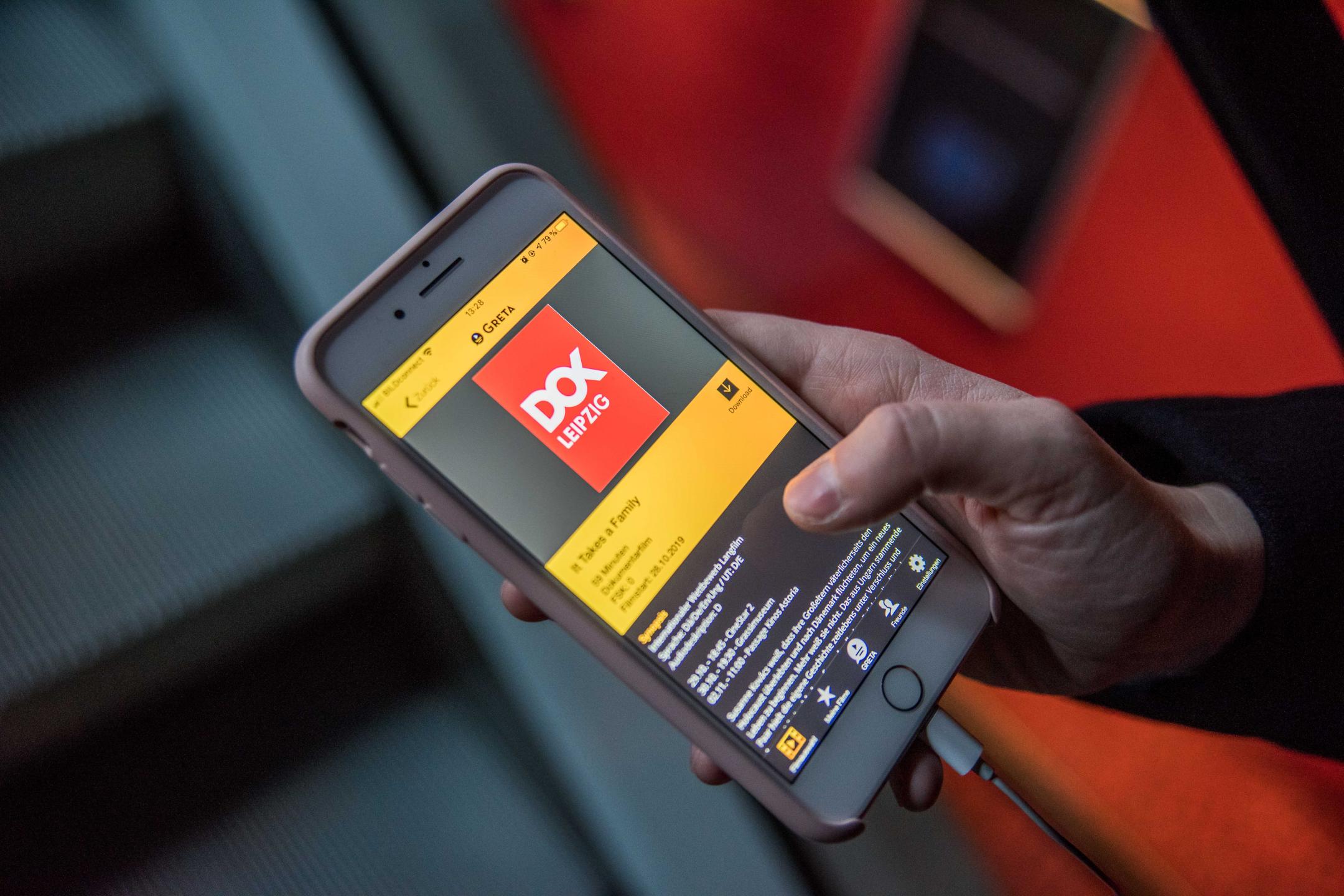 Eine Besucherin, von der nur die rechte Jand zu sehen ist, hält darin ihr Smartphone. Auf dem Display erscheint die App Greta, ausgewählt ist der Film It Takes a Family.