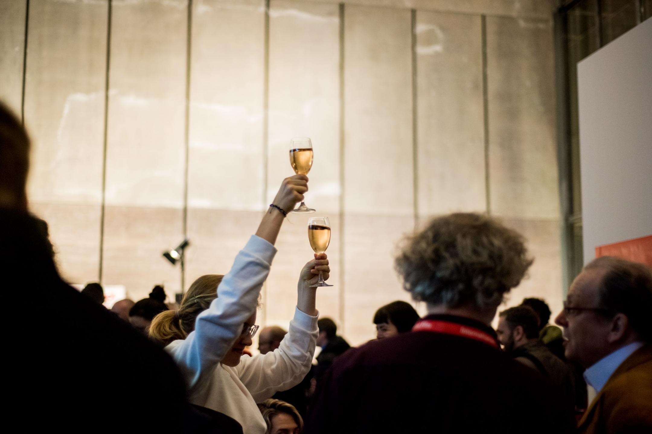 A woman with two glasses of wine in the middle of a crowd.