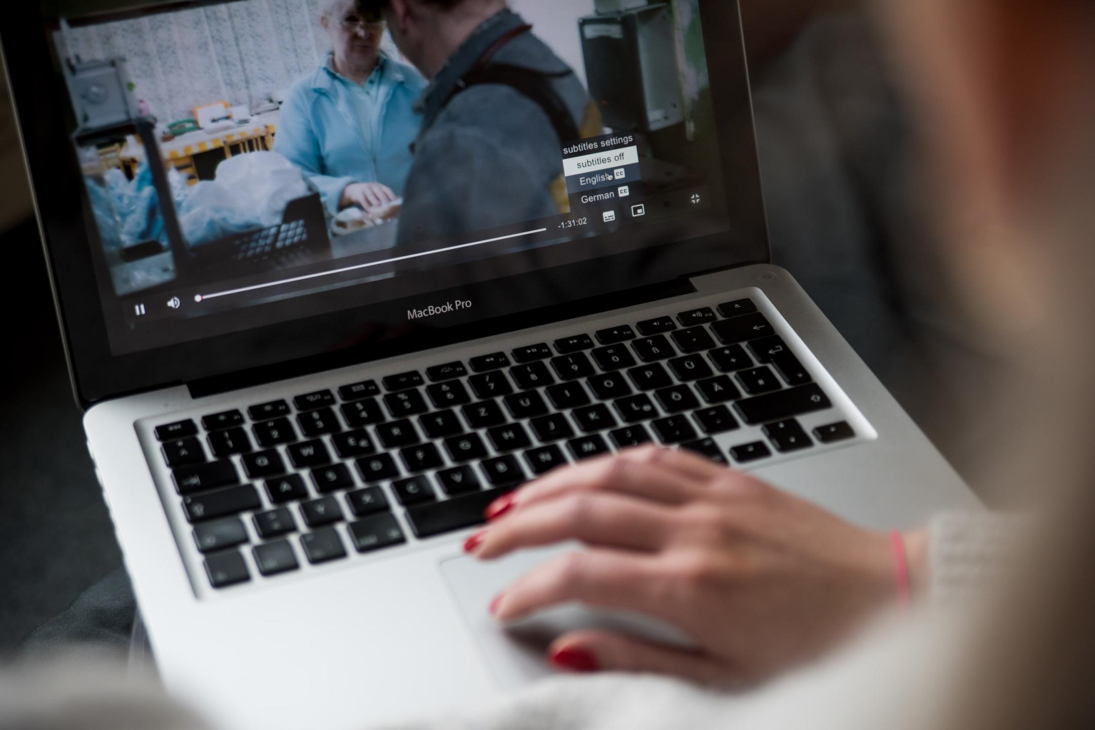Eine Hand bedient das Mauspad eines MacBooks, auf dem Laptop-Bildschirm ist ein Videomenü aufgeklappt mit den Wahloptionen Subtitles off, English und German.