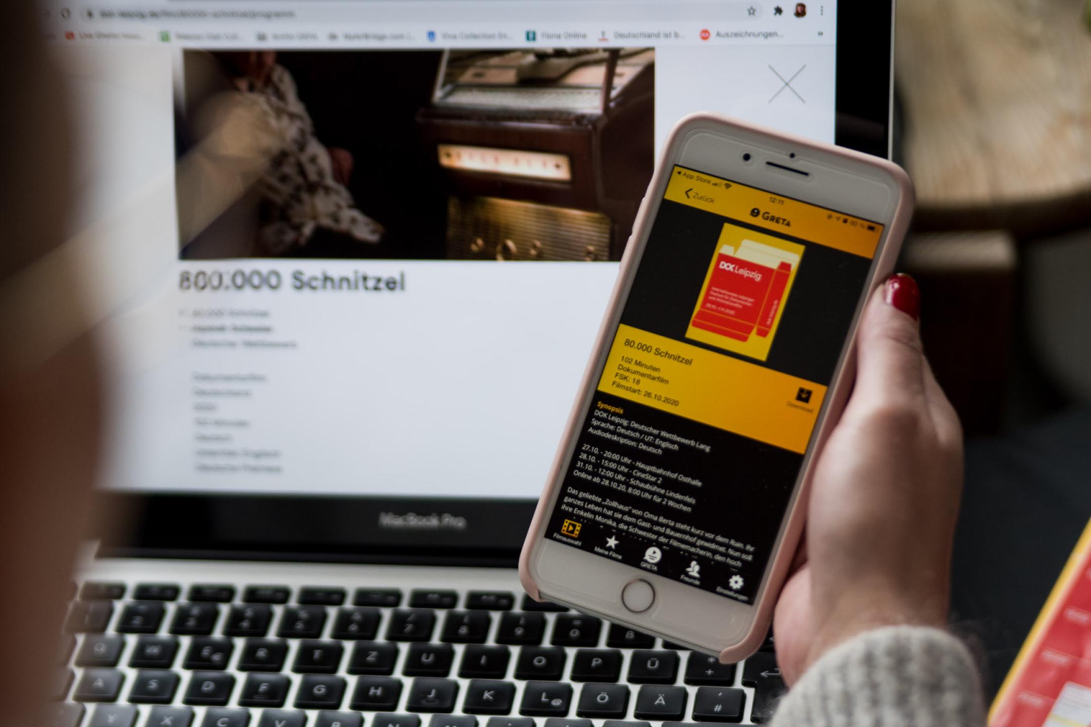 Blick über die Schulter einer Person auf ihren Laptopbildschirm, in der Hand hält sie ein Smartphone mit der App Greta.
