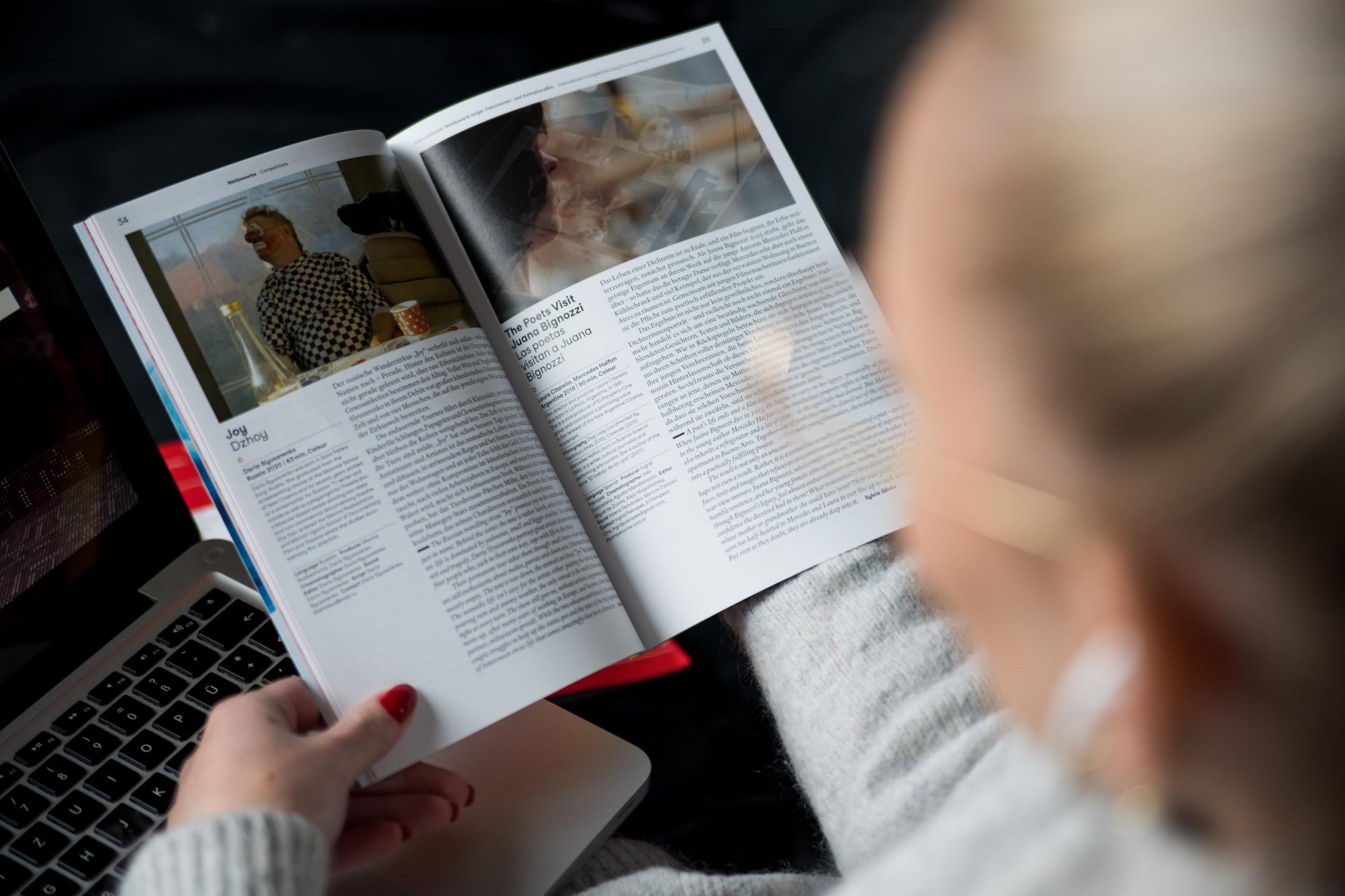 Eine Frau blättert im DOK Festivalkatalog von 2021, die Kameraperspektive blickt über ihre Schulter auf den Katalog in ihrer Hand.