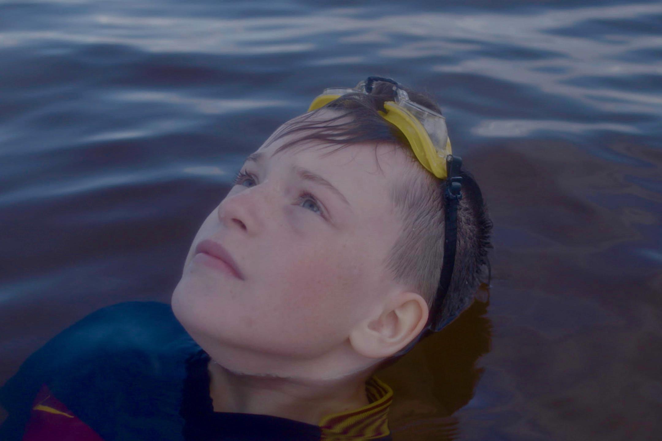 Filmstill aus "Gabi": Ein Junge mit Schwimmbrille auf dem Kopf liegt im Wasser und blickt in den Himmel.