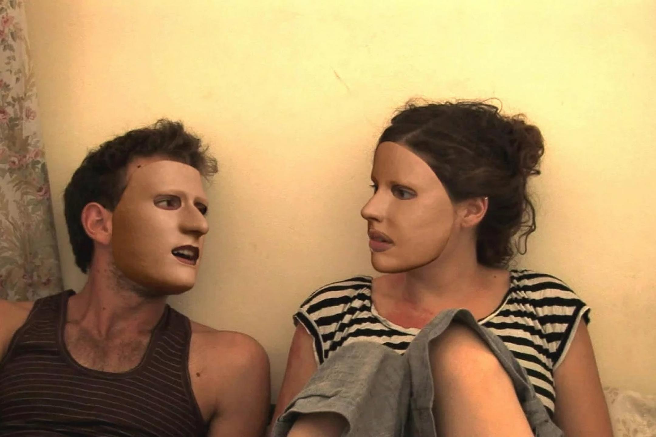 Filmszene aus Z32: Ein Mann und eine Frau sitzen nebeneinander an eine weiße Wand gelegt. Ihre Köpfe sind einander zugewandt. Beide tragen Masken, die ihr ganzes Gesicht überdecken.