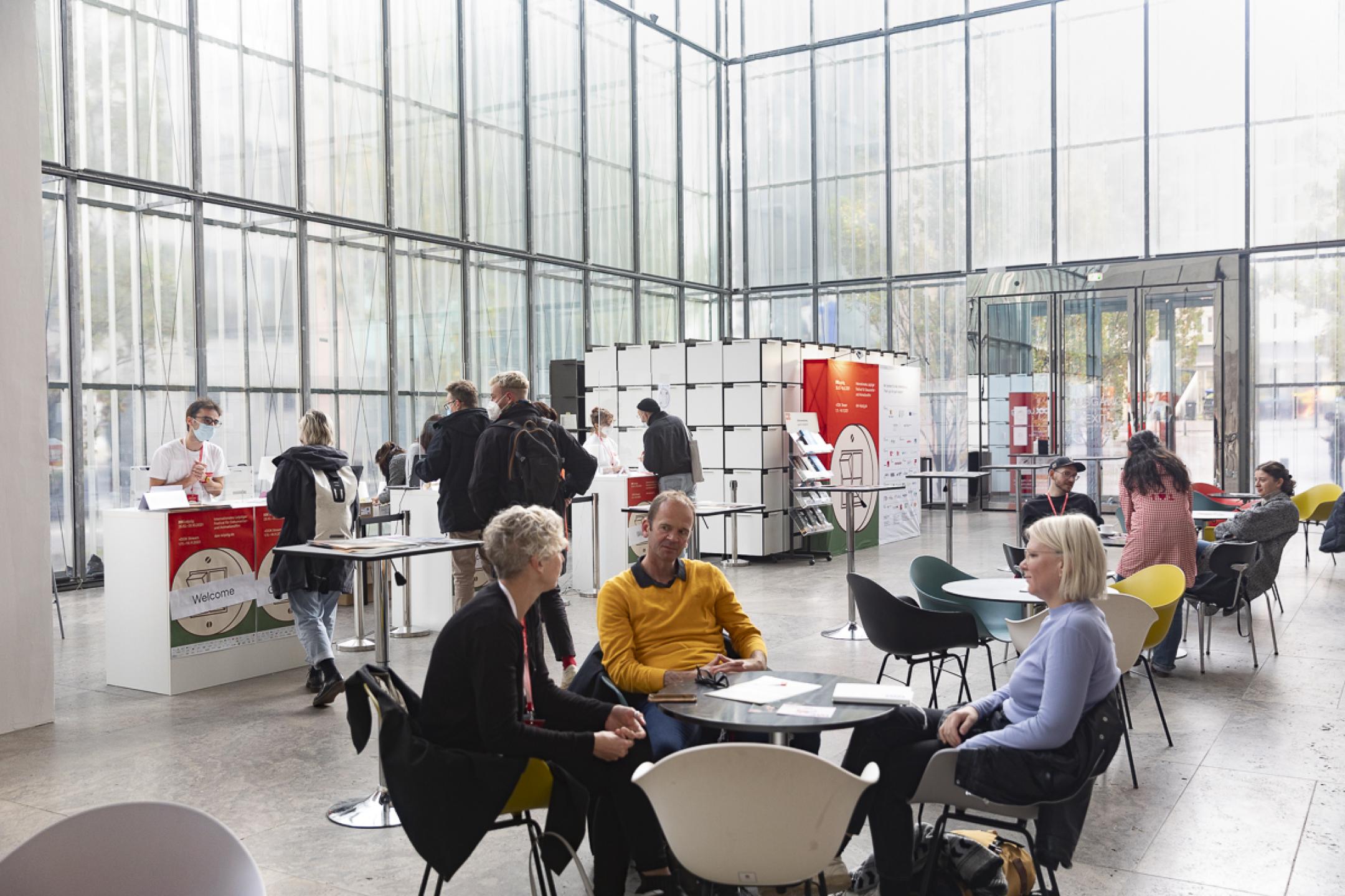 Blick in das Festivalzentrum von DOK Leipzig: Ein hoher Raum mit gläsernen Wänden, darin mehrere Personen im Gespräch an Kaffeetischen, im Hintergrund der Gästecounter mit mehreren Personen, die dort anstehen.