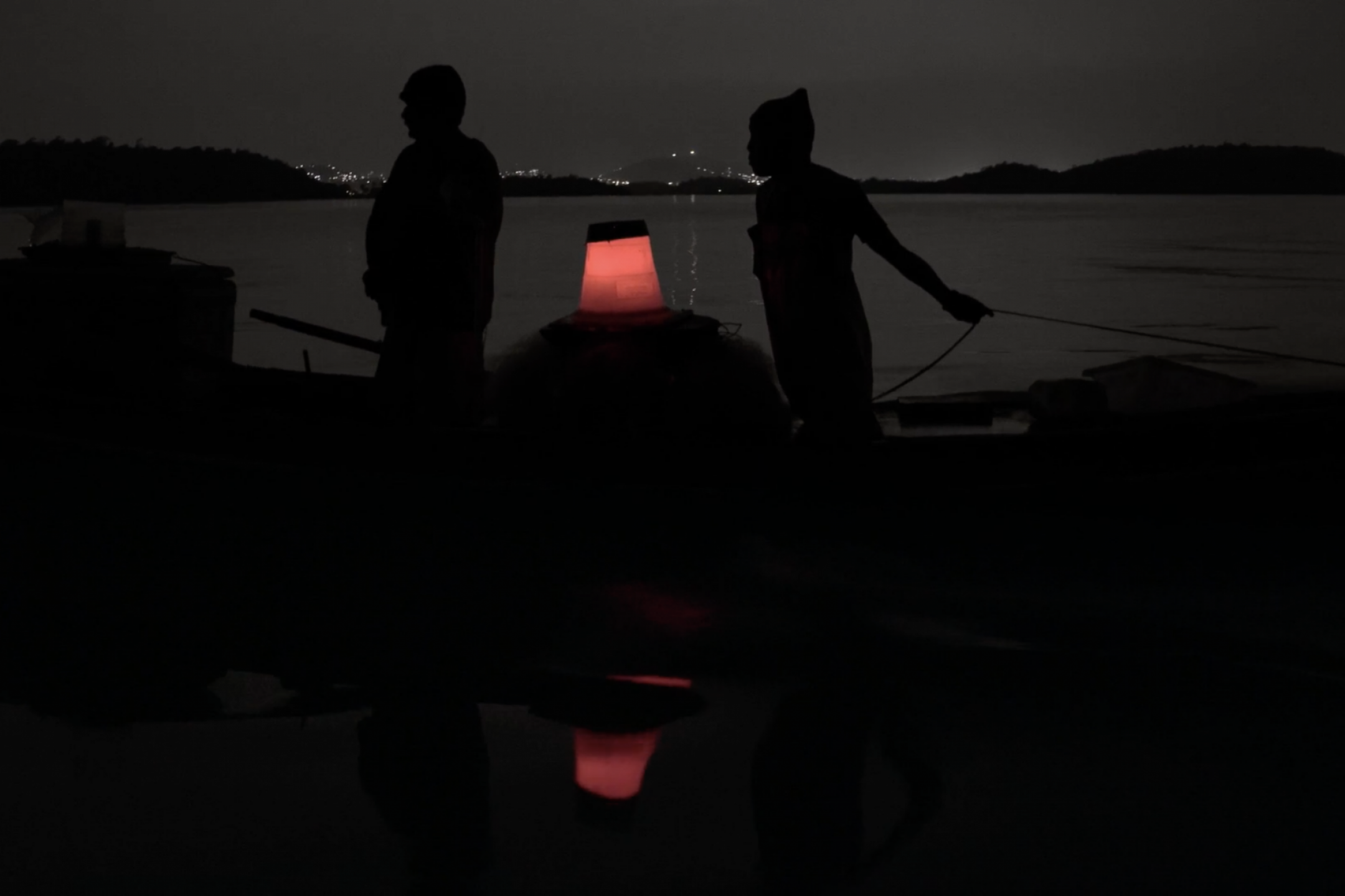 A night scene: Two persons are standing on a small boat, in the background the shapes of a bay are visible. The only light is a small lantern on the boat.