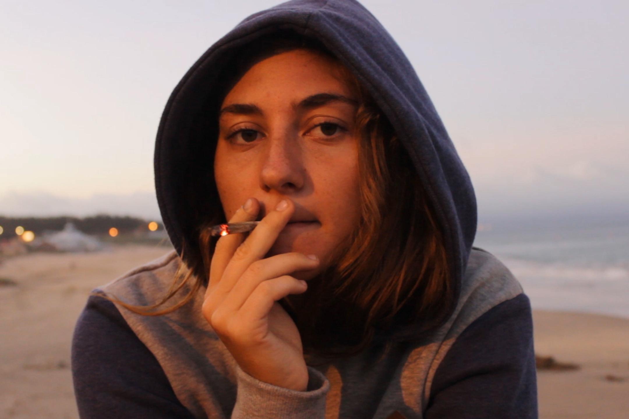 Porträt einer jungen Frau in einem Hoodie mit Kapuze. Sie raucht eine Zigarette und blickt lässig-nachdenklich in die Kamera.