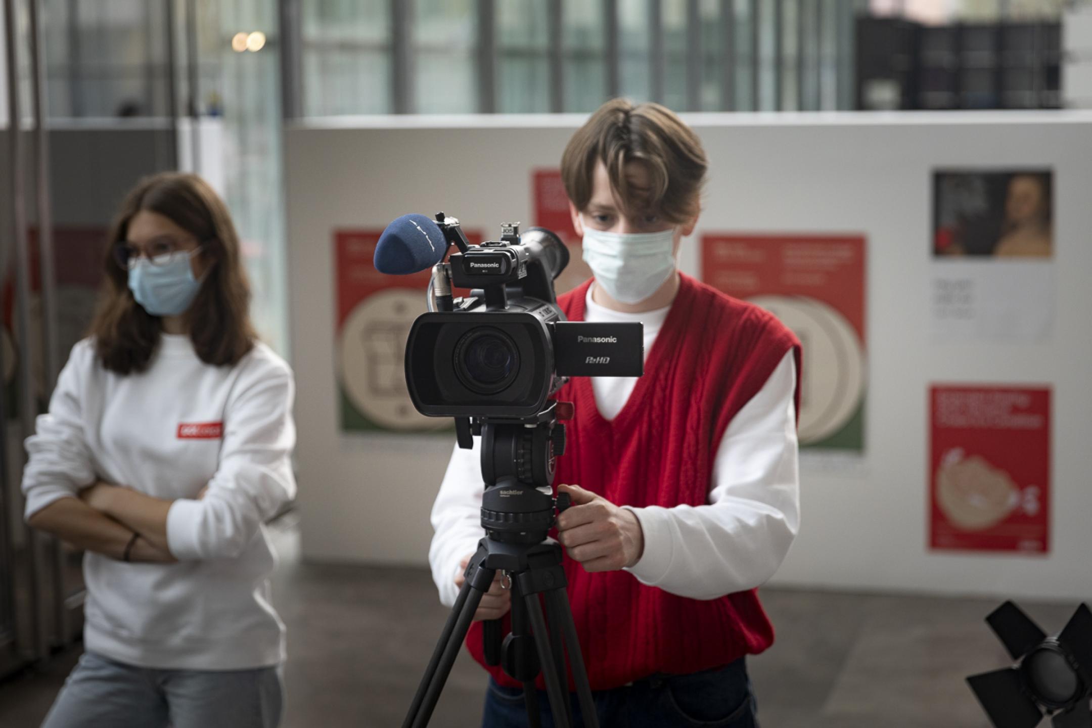 Ein Jugendlicher positioniert eine semi-professionelle Kamera mit Aufsteckmikrofon und Stativ im Festivalzentrum von DOK Leipzig. Im Hintergrund mehrere DOK-Plakate.