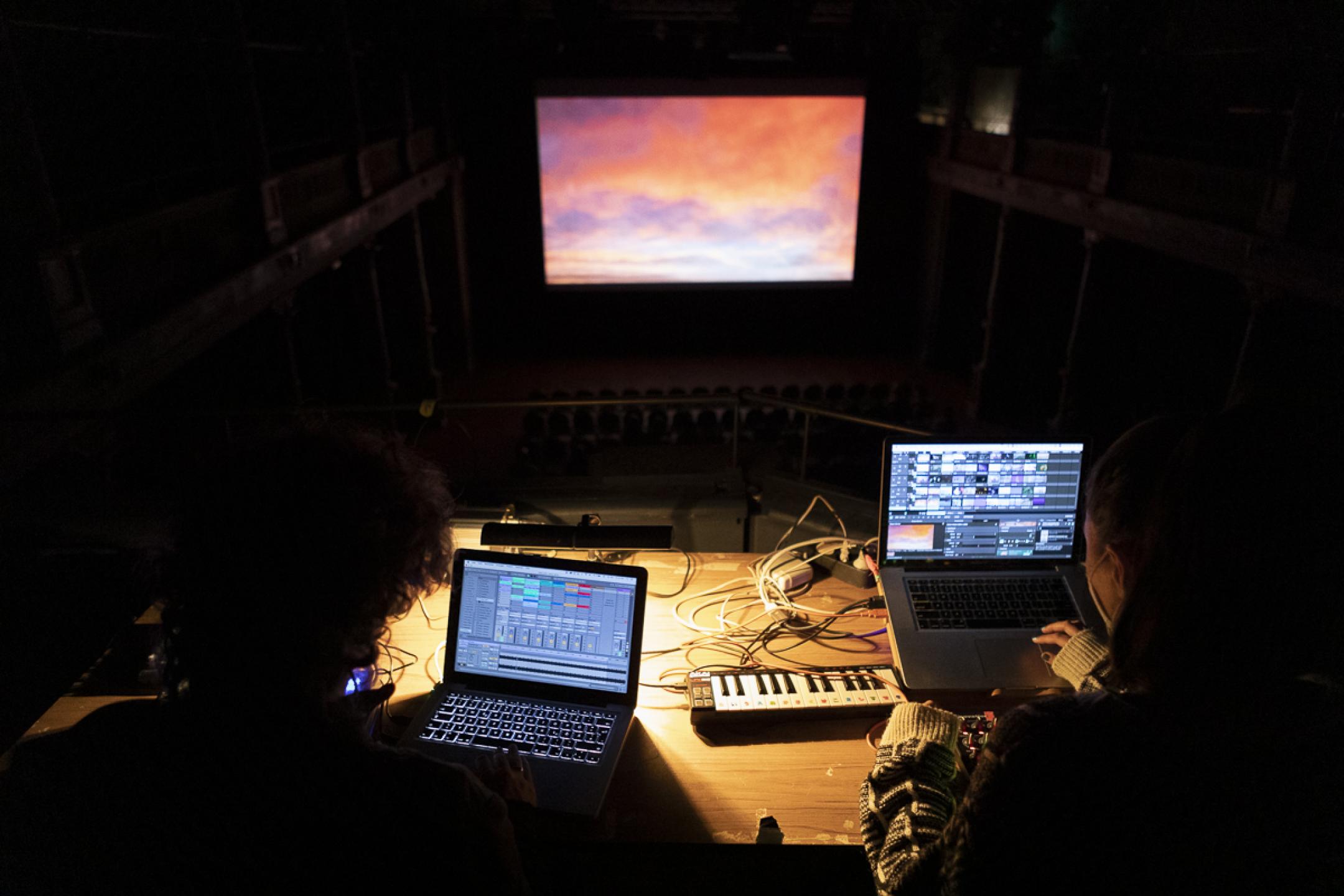 Die beiden Künstlerinnen CFM und SAOU TV sitzen auf einer Technik-Empore in einem dunklen Kinosaal. Sie sammeln Bilder und Sounds an ihren beiden Laptops. Im Hintergrund zeigt die erleuchtete Leinwand Wolkenbilder im Abendrot.