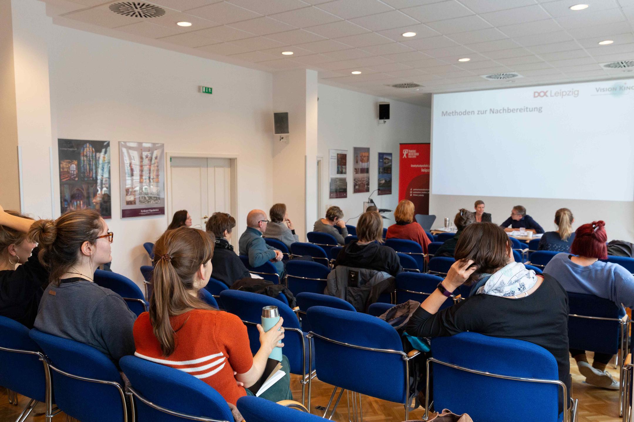 Ein Veranstaltungsraum, in dem verteilt mit Mindestabstand mehrere Personen sitzen und drei Vortragenden zuhören. Auf der Leinwand hinter den Vortragenden steht der Schriftzug: Methoden der Nachbereitung. 