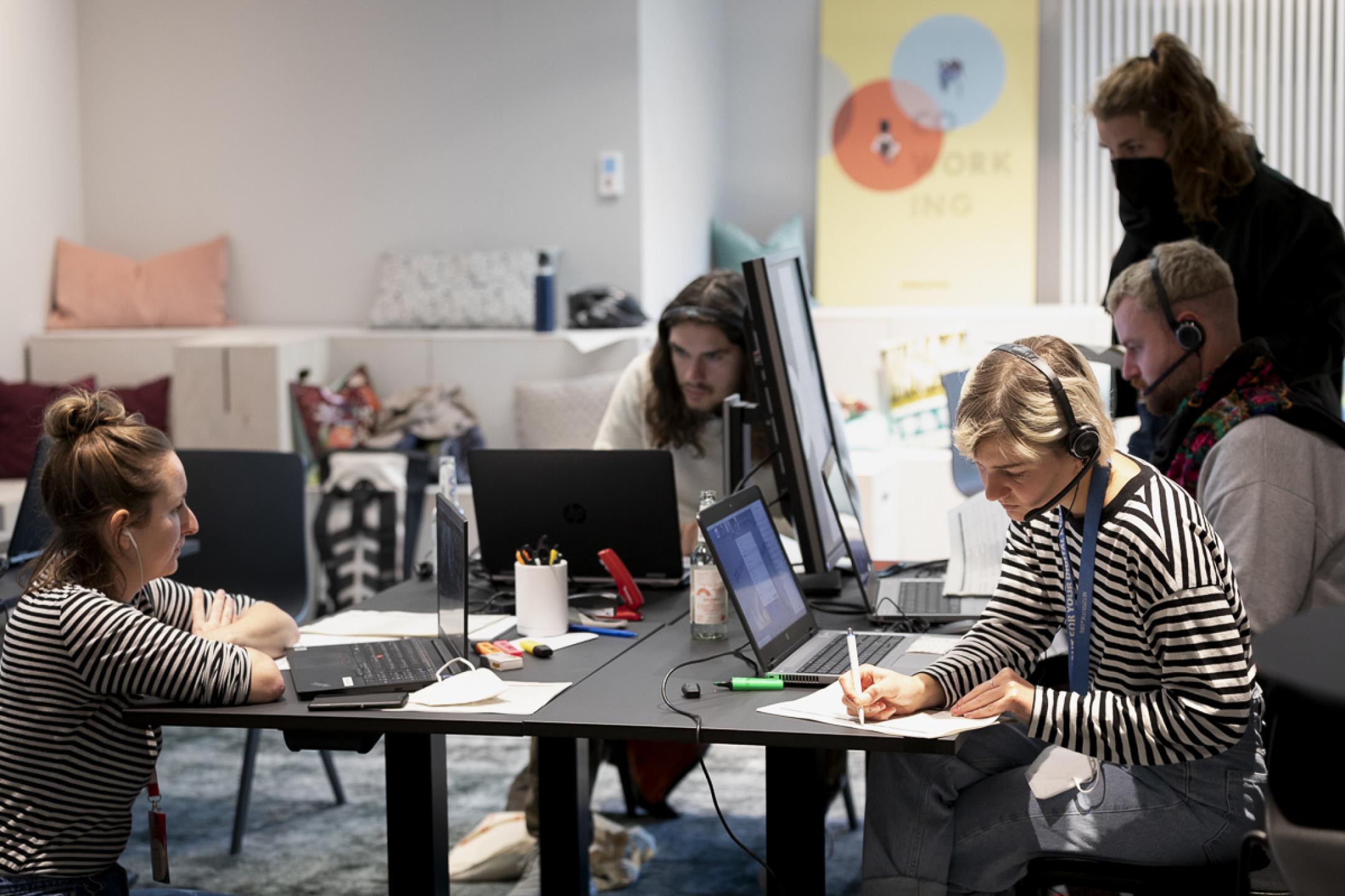 Fünf Personen gruppieren sich sitzend und stehend um einen großen quadratischen Arbeitstisch mit mehreren Laptops. Eine Frau (rechts im Bild) macht sich  Notizen auf einen DIN A4-Zettel.