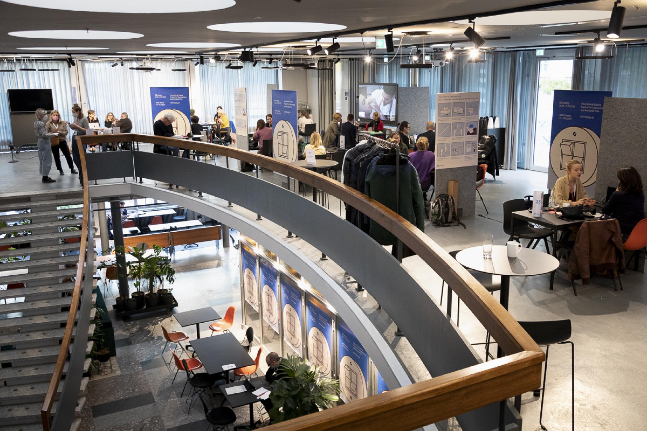 Overview of the Co-Pro Market meetings: A huge staircase is leading to the second floor of an open office space. There are several desks in open meeting compartments. Smalls groups of people (2–4) are sitting at the desks. 