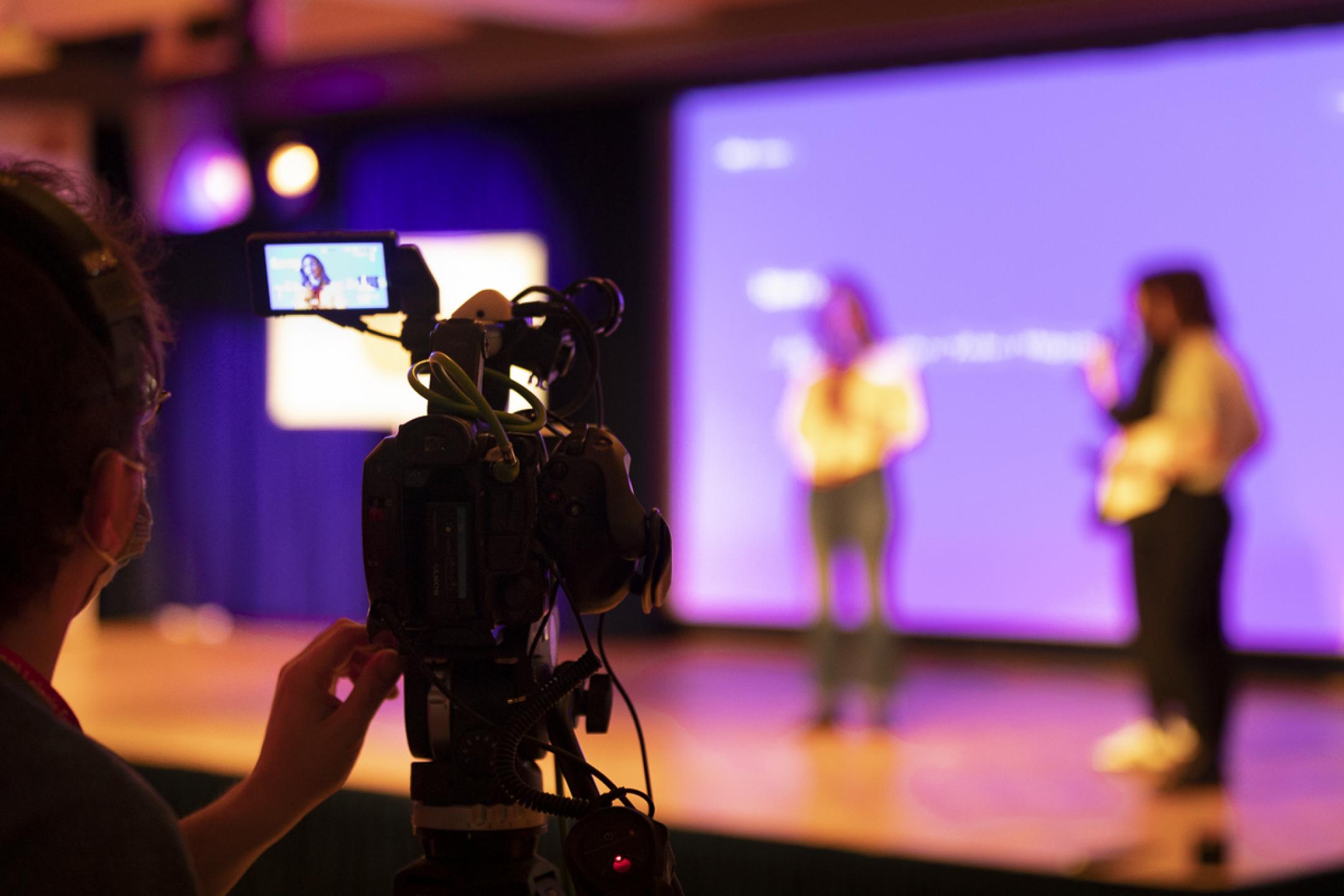 A tripod camera in the foreground. In the background, three people present on an event stage. 