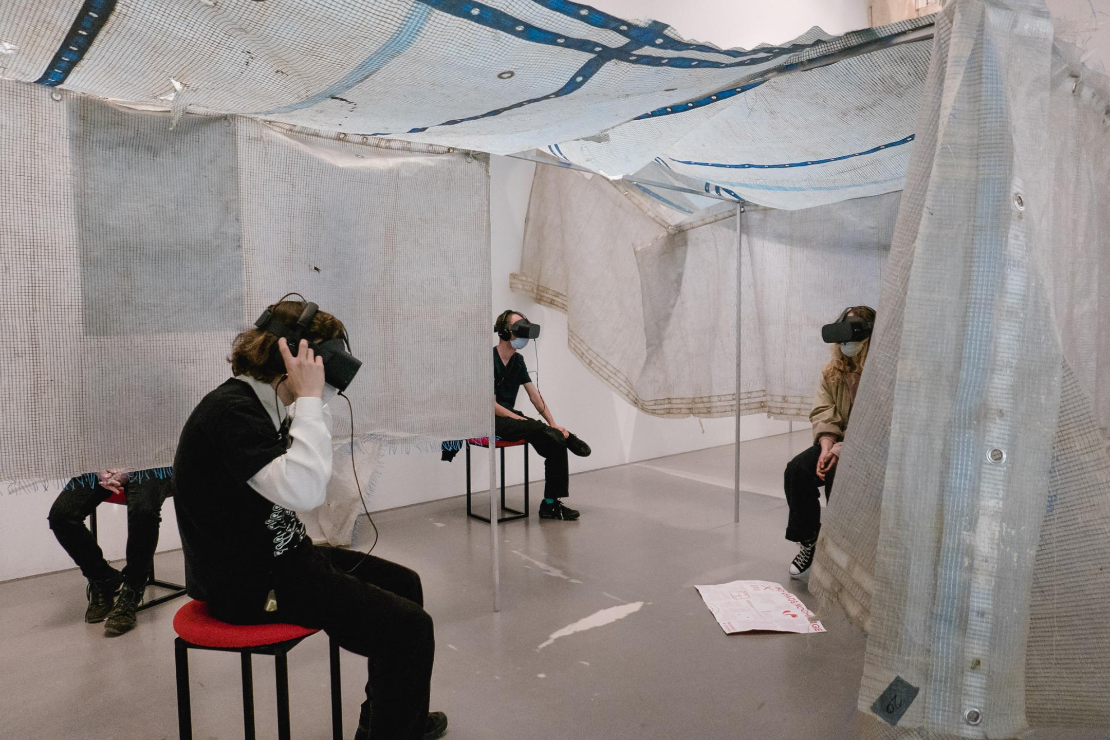 Ein riesiges Zelt aus heller Meshplane, das durch herabhängende Teile in einzelne Räume geteilt wird. Vier Personen mit VR-Brillen sitzen in verschiedenen Ecken des Zeltes auf Hockern.