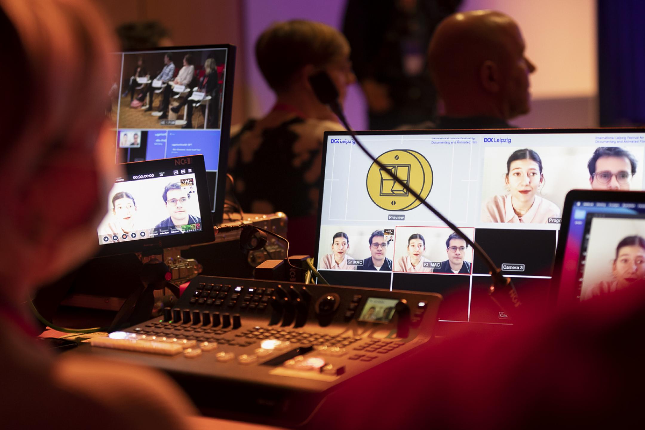 A live editing suite at a hybrid event: A man is sitting surrounded by four monitors of different sizes, showing the event stage and people waiting in zoom.
