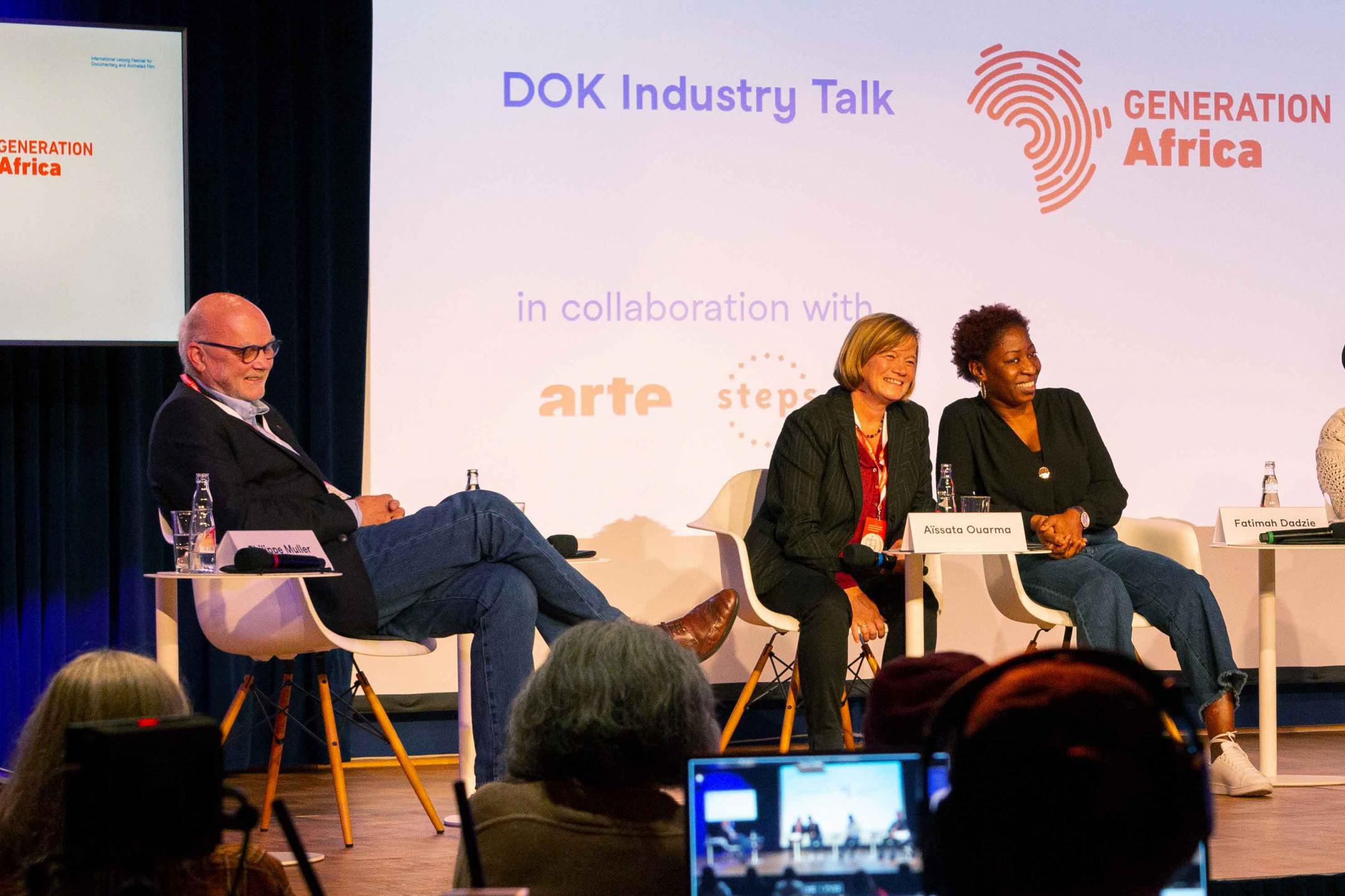 Three persons on stage of a panel, all of them laughing and looking to the audience. FLTR: A man in blue jeans, a woman with blond hair who is wearing a black suit, and a black woman with short hair. In the background a big screen with information on the event: DOK Industry Talk, below the logos of arte and steps.