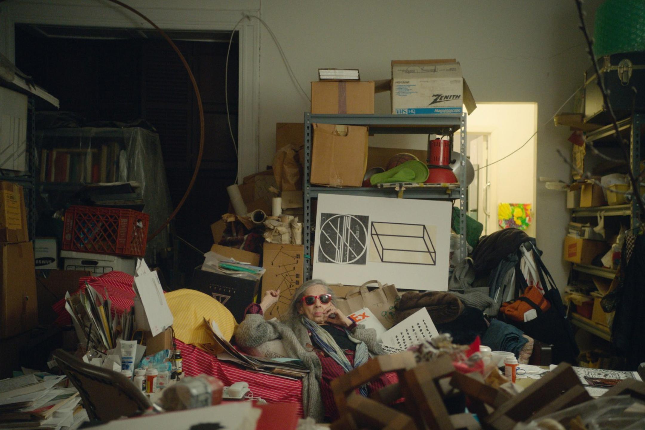 An elderly woman wearing sunglasses sits on the floor of a storage room that is fully packed with boxes, bags and furniture