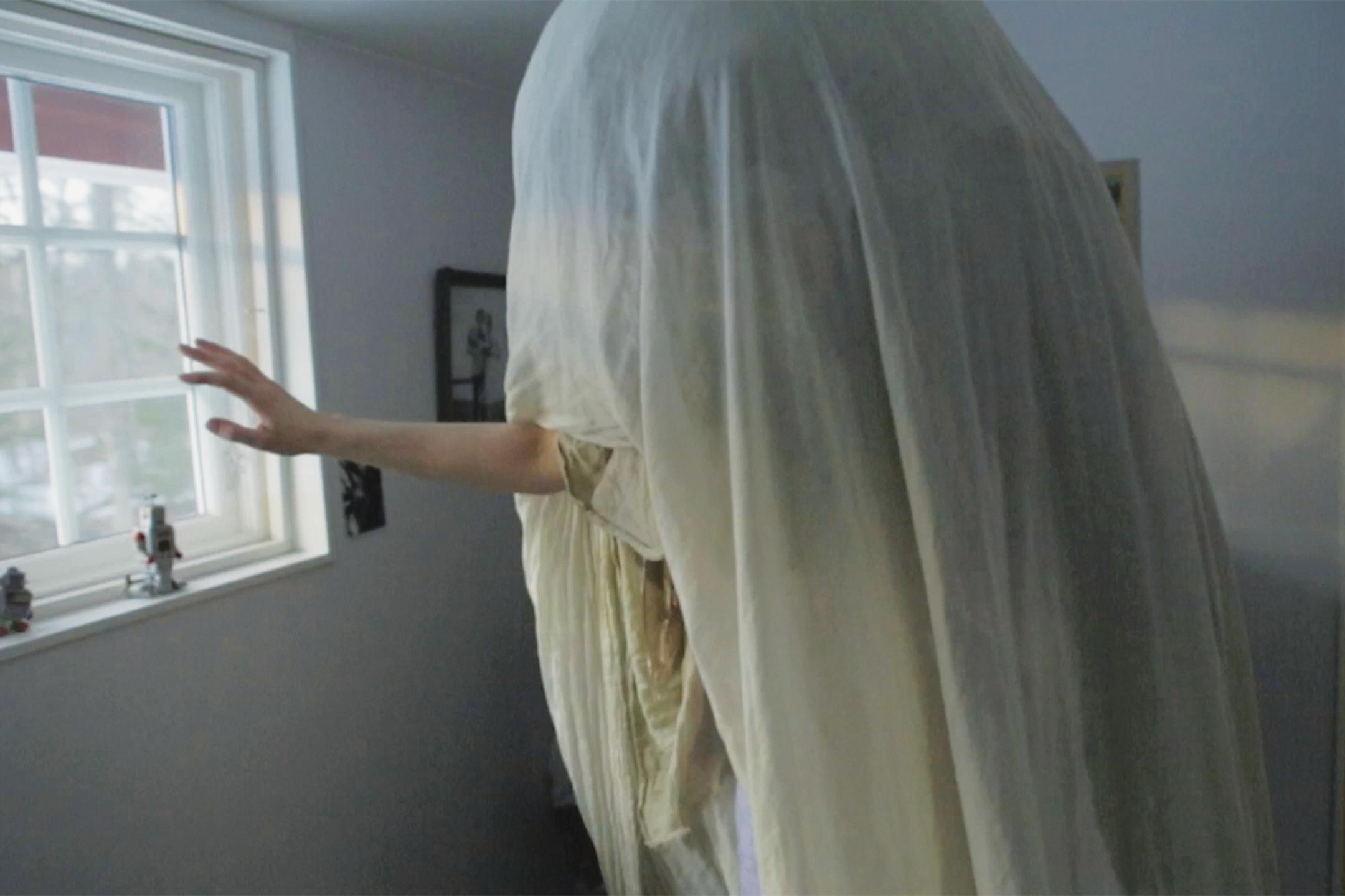 A person wrapped in many layers of white fabric stands in the middle of a living room. Only the persons arms is visible, pointing out of the fabric in direction of the window.