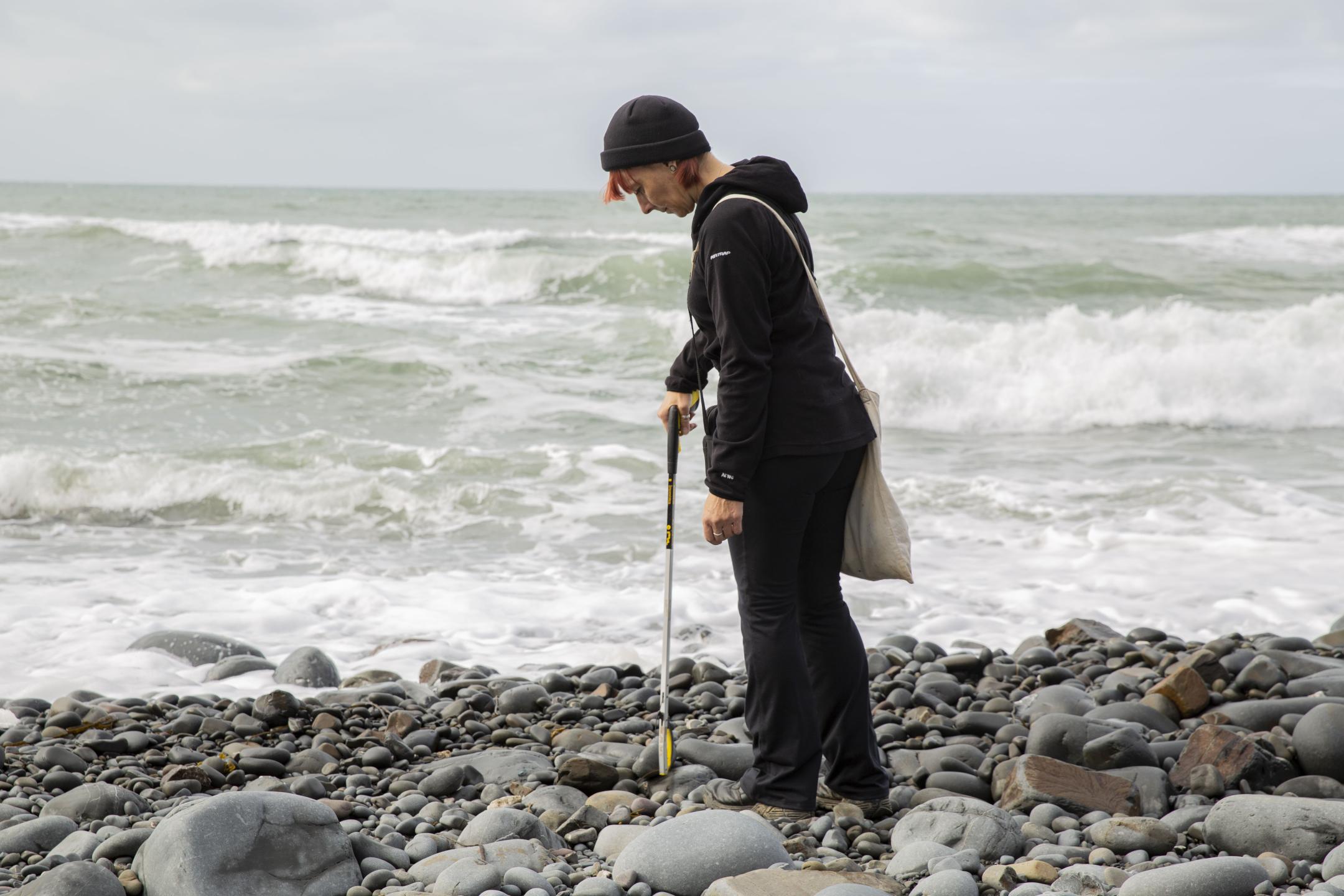 A woman is walking at a rocky beach. She looks down and picks something up with a long gripper.