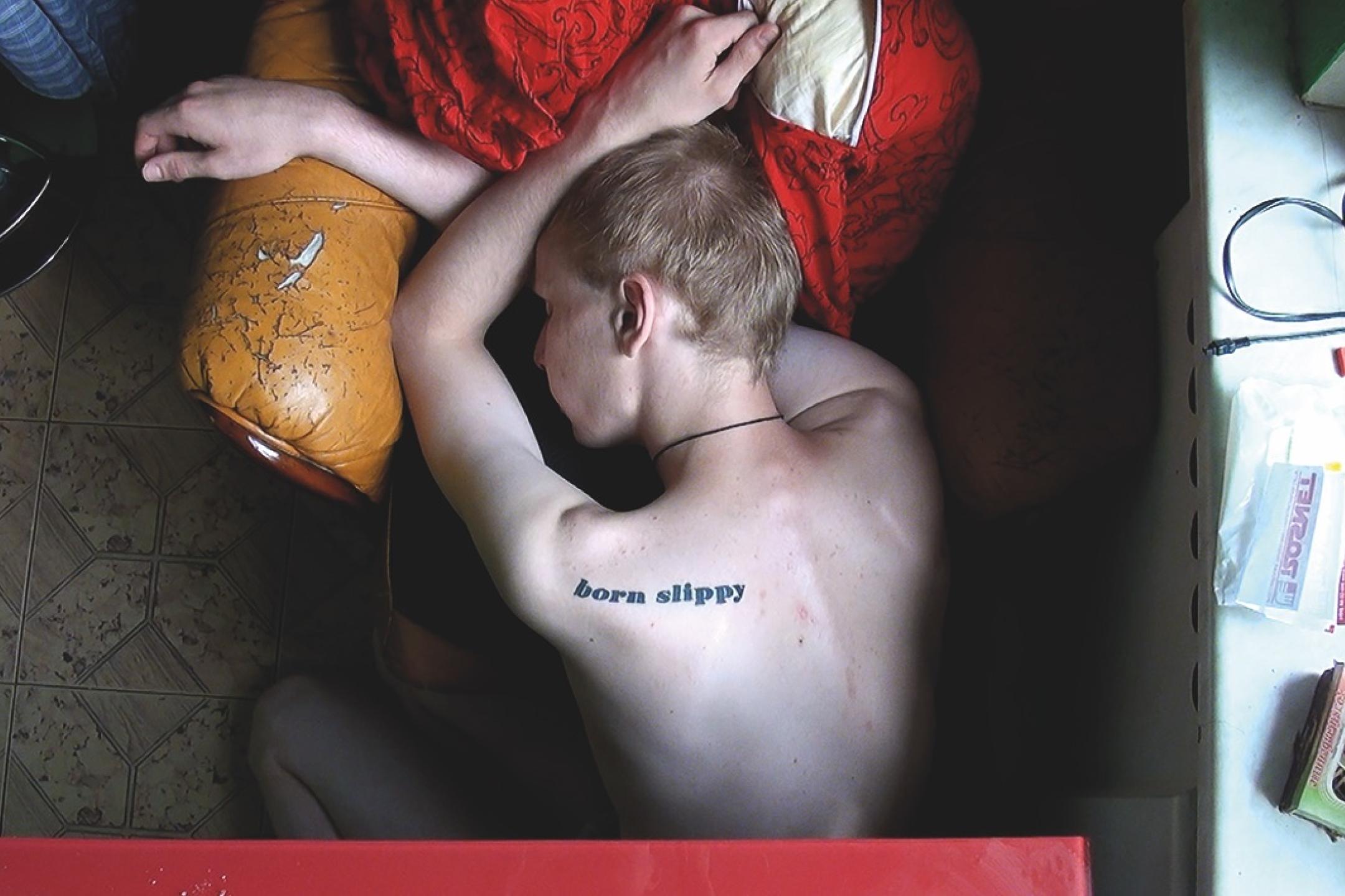From a bird’s eye view: A male teenager is resting his head on a chair. He does not wear a shirt. On his shoulder, you can see a tattoo of the words "Born slippy".