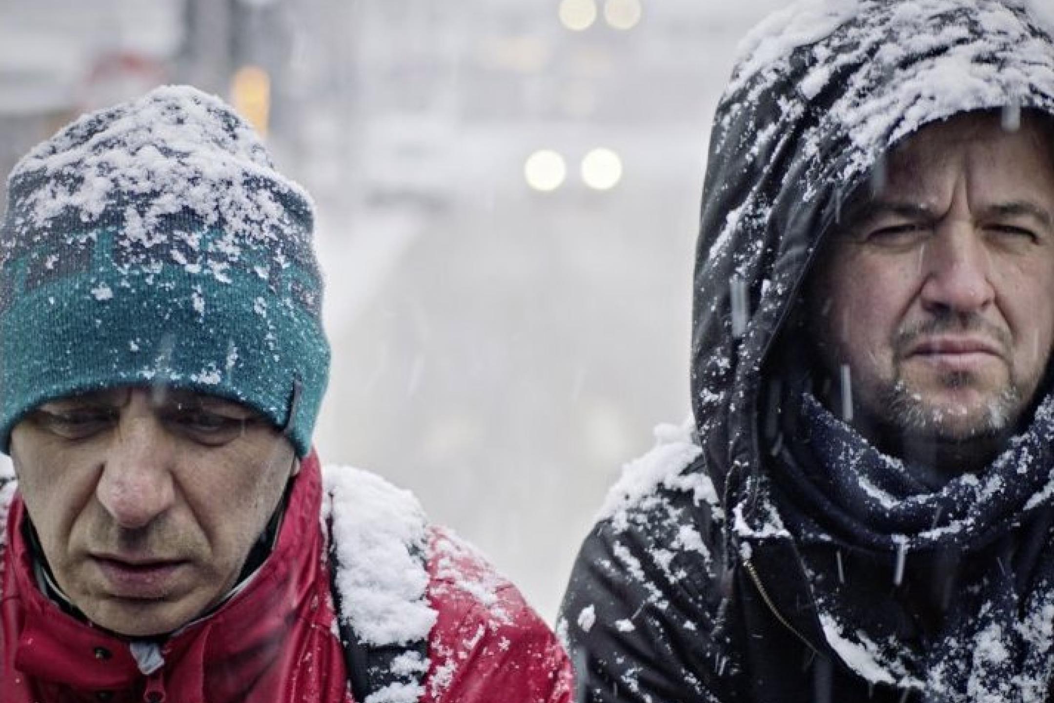 Two grim-looking men who are wearing winter jackets covered with snow are walking side by side in a street that is also covered with snow.
