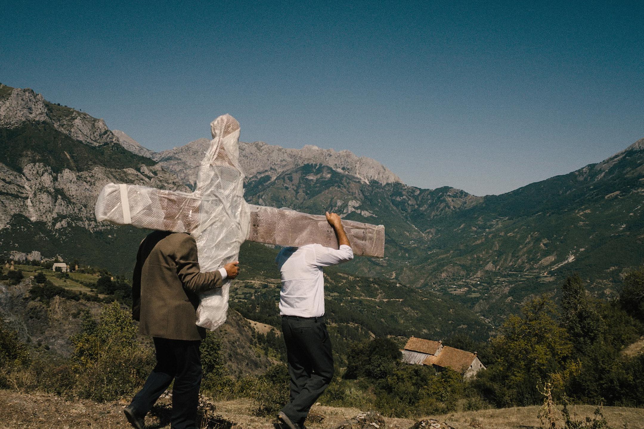 Amidst the scenery of alpine mountains, three men are walking down a path from a hill. They  carrying a huge cross on their shoulders.