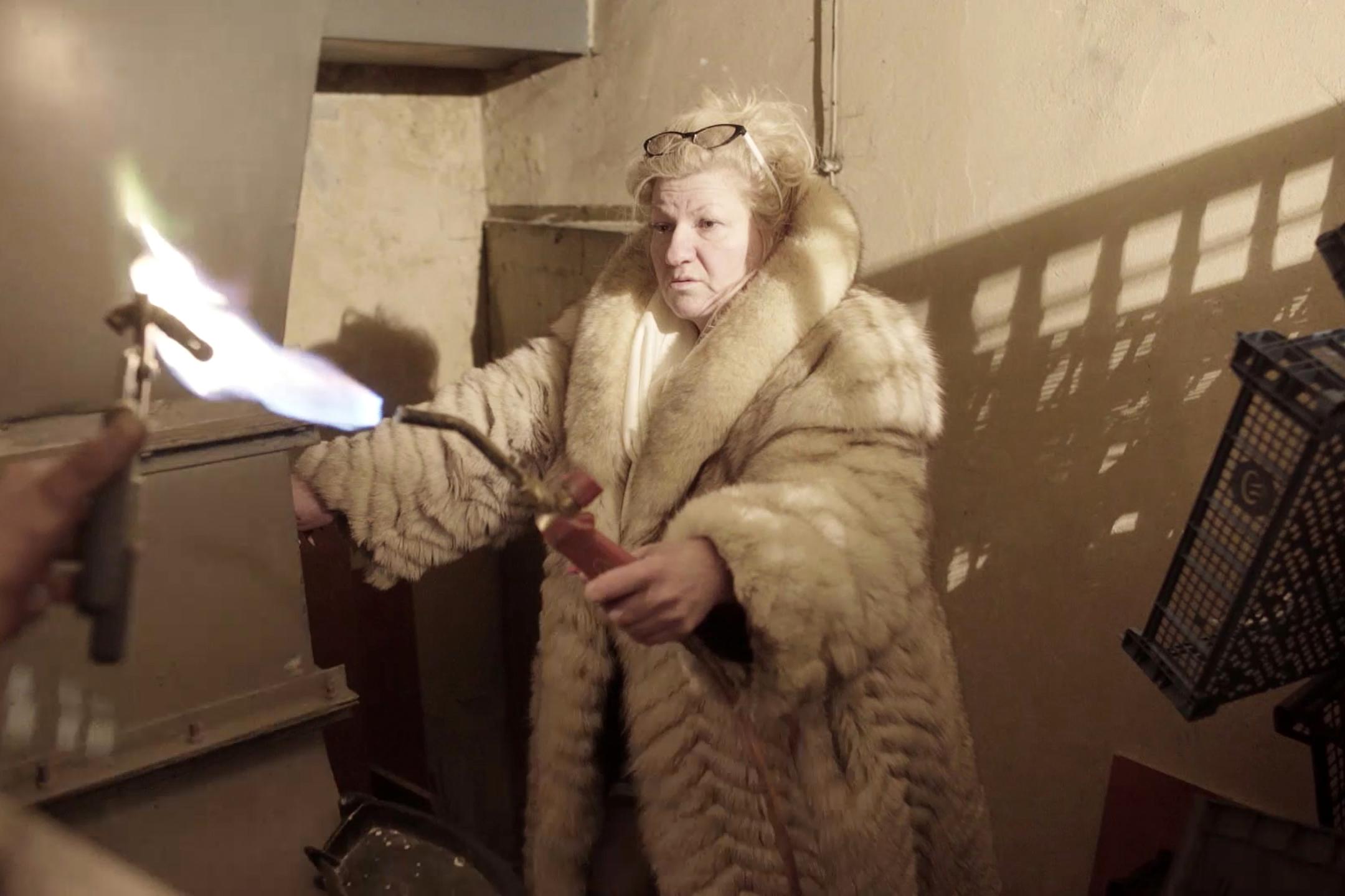 A woman with a fur coat holds a Bunsen burner with a huge flame.