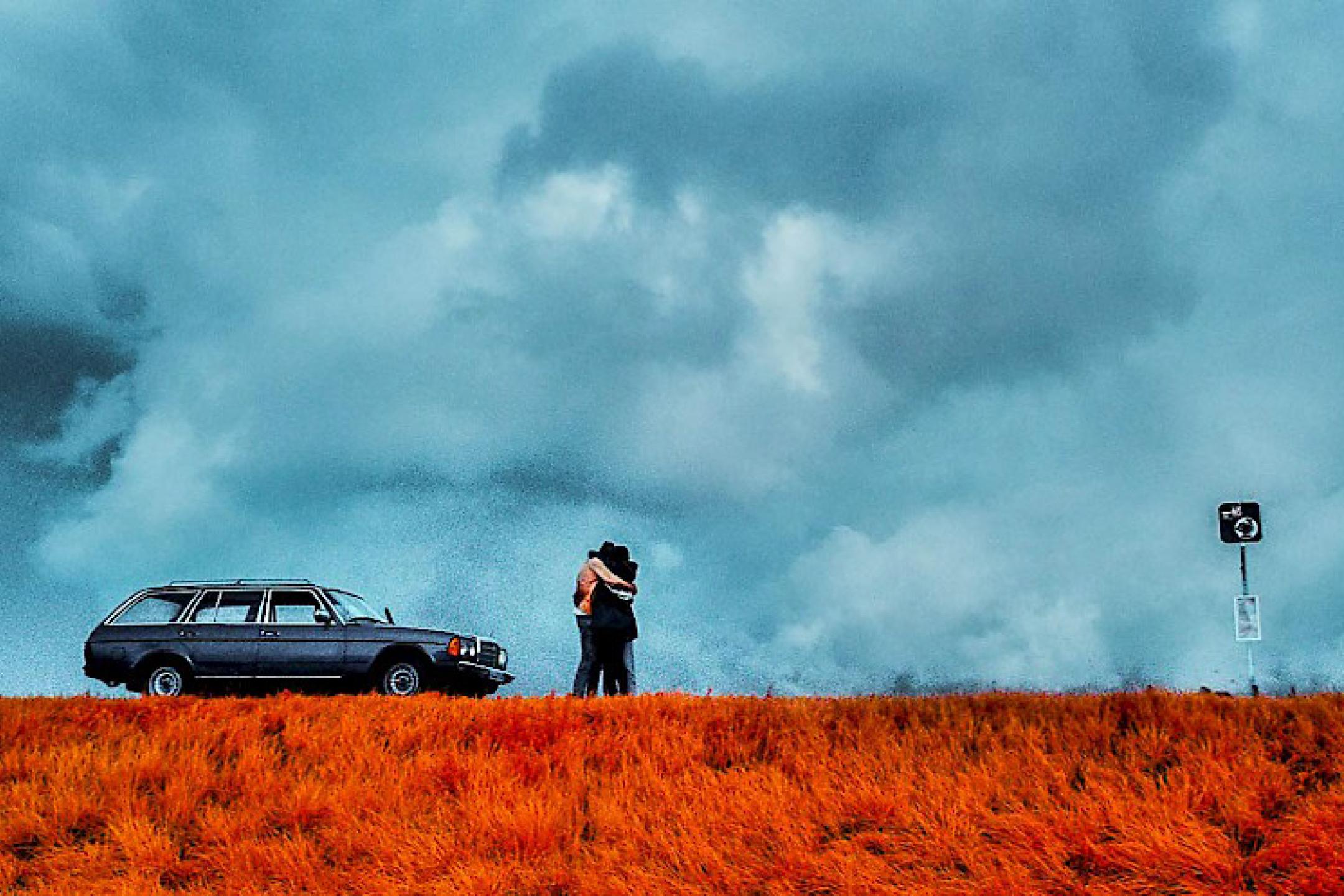 A wide corn field under a thunderstorm sky. A couple stands in the field, hugging each other. On the side parks a car.