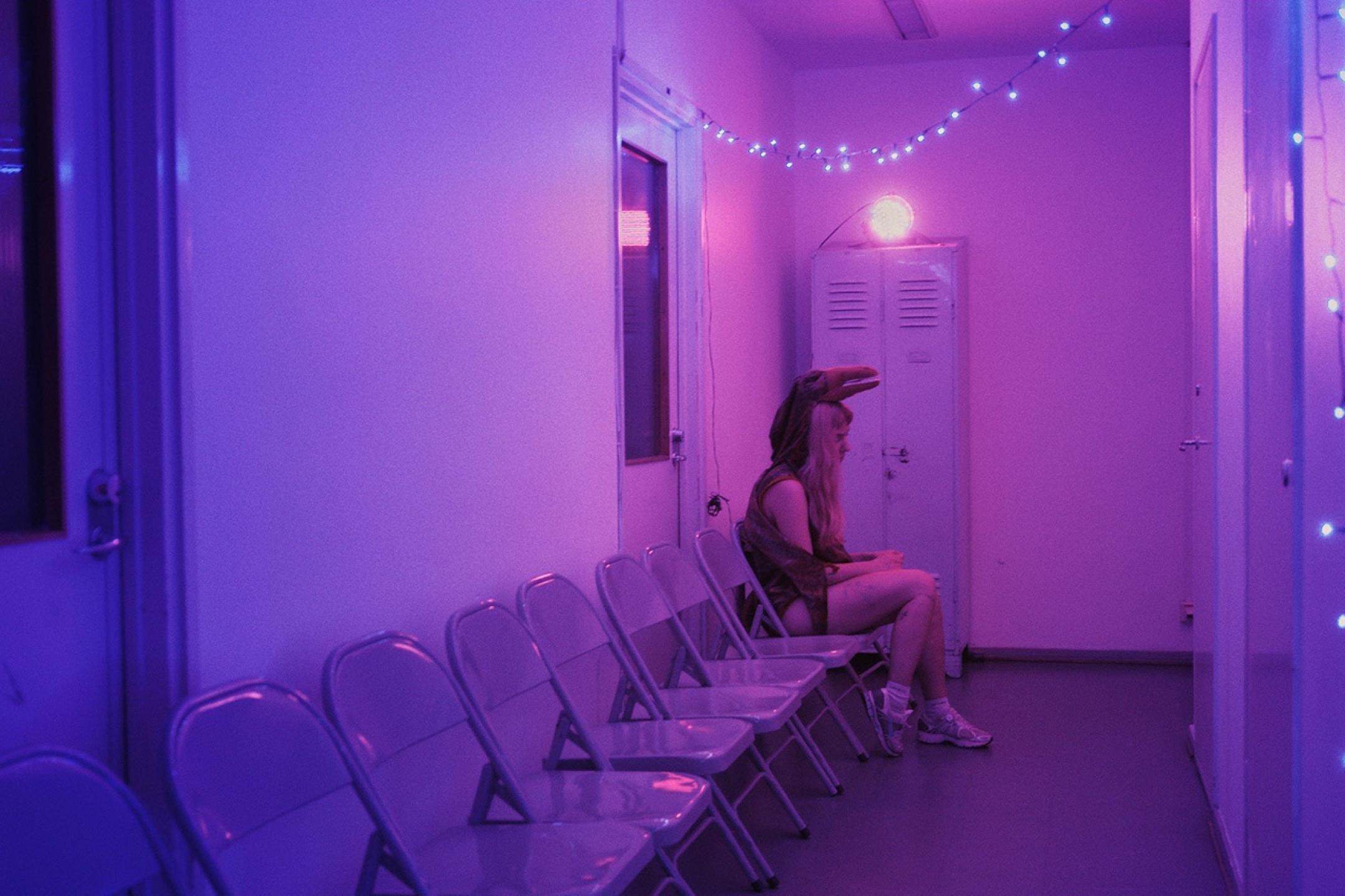 A woman in a bird costume is sitting on a plastic chair, all alone in a very clean waiting room. Above her head a small chain of lights that creates a purple lighting mood.
