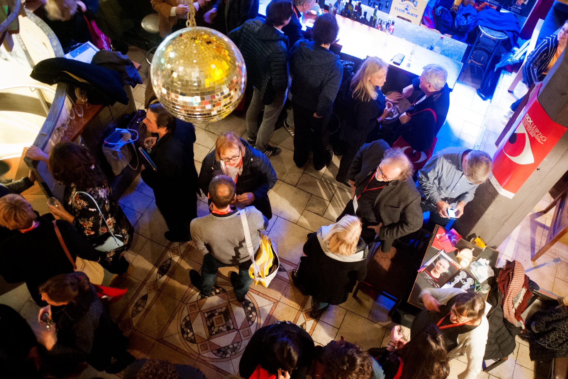 A business cocktail hour from a bird’s eye view: about 20 people standing in small groups and chatting, some are holding glasses, some are ordering at the bar. 