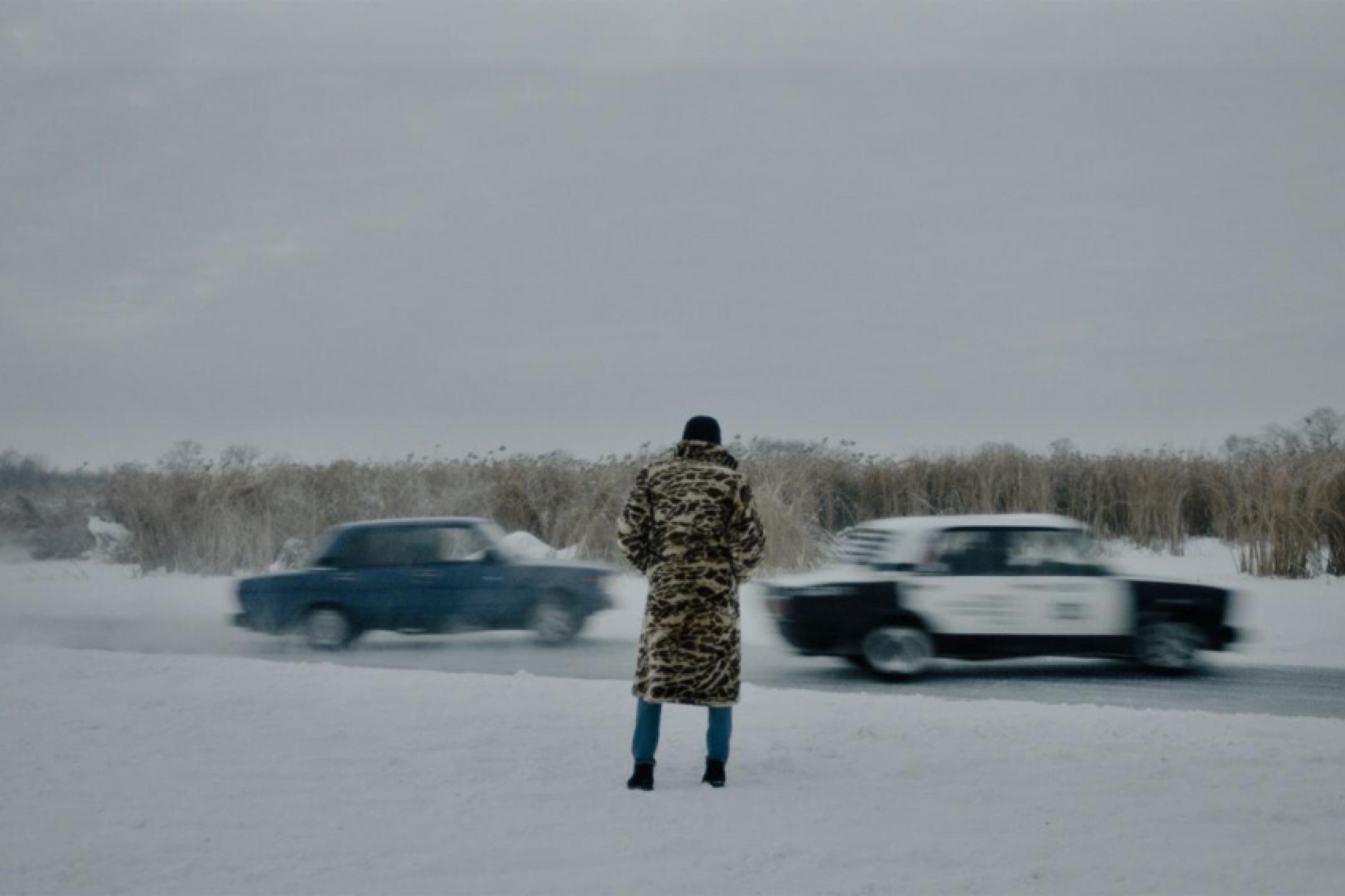 A street covered with snow in winter: A man with a leopard fur coat is watching two cars racing through the snow.