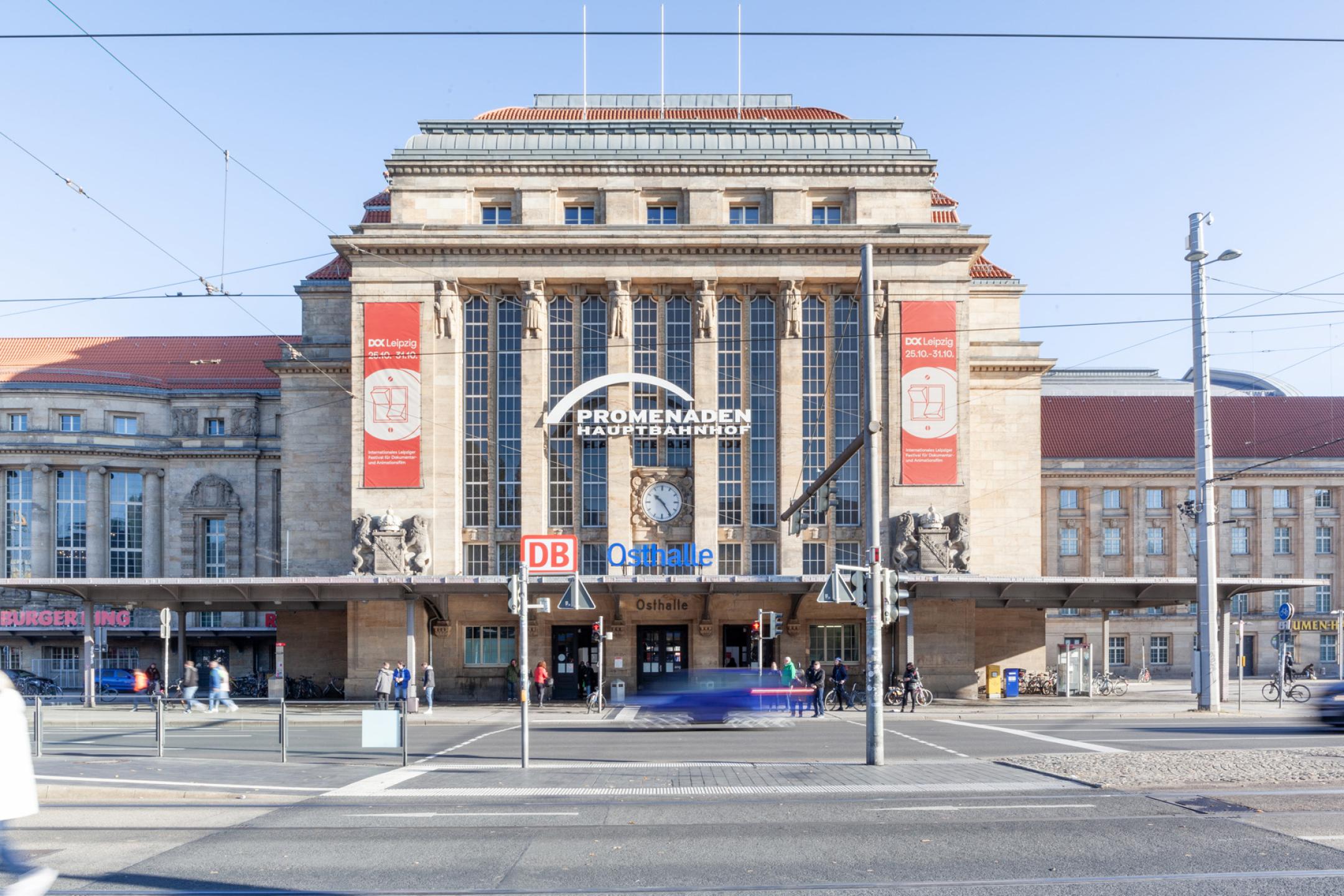Blick auf den Vorplatz und die Osthalle des Leipziger Hauptbahnhofs. Links und rechts neben dem Eingang  zwei rote Banner von DOK Leipzig.