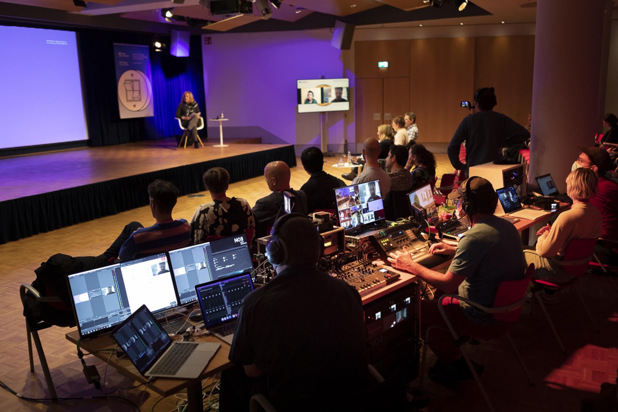 An event space: A woman sits in a chair on stage. Several people are listening in the first row. Behind them a huge live editing unit with eight screens, operated by two editors who are wearing headphones.
